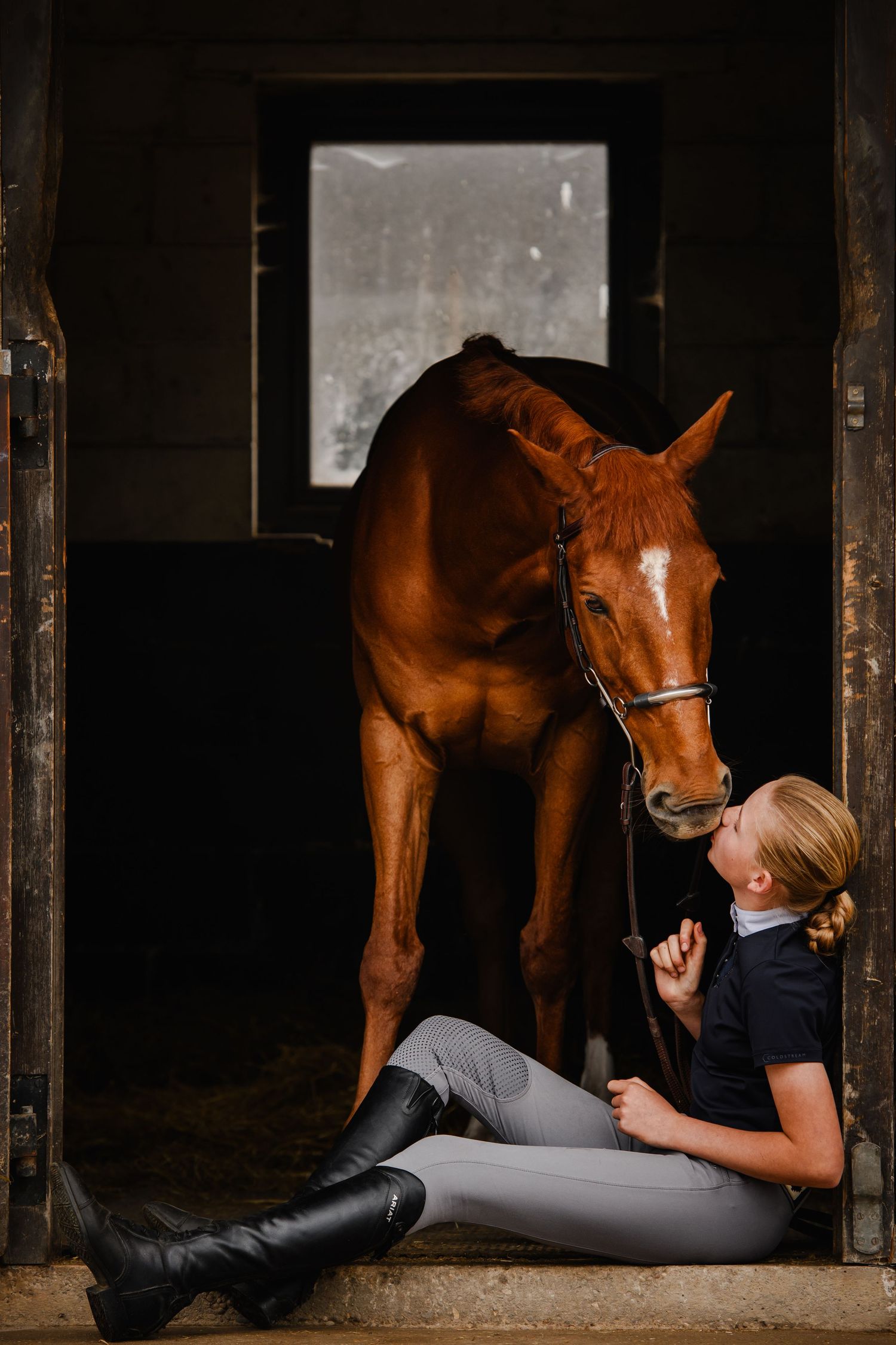 A young female equestrian sits on stable floor bonding with a chestnut horse in riding boots and breeches.