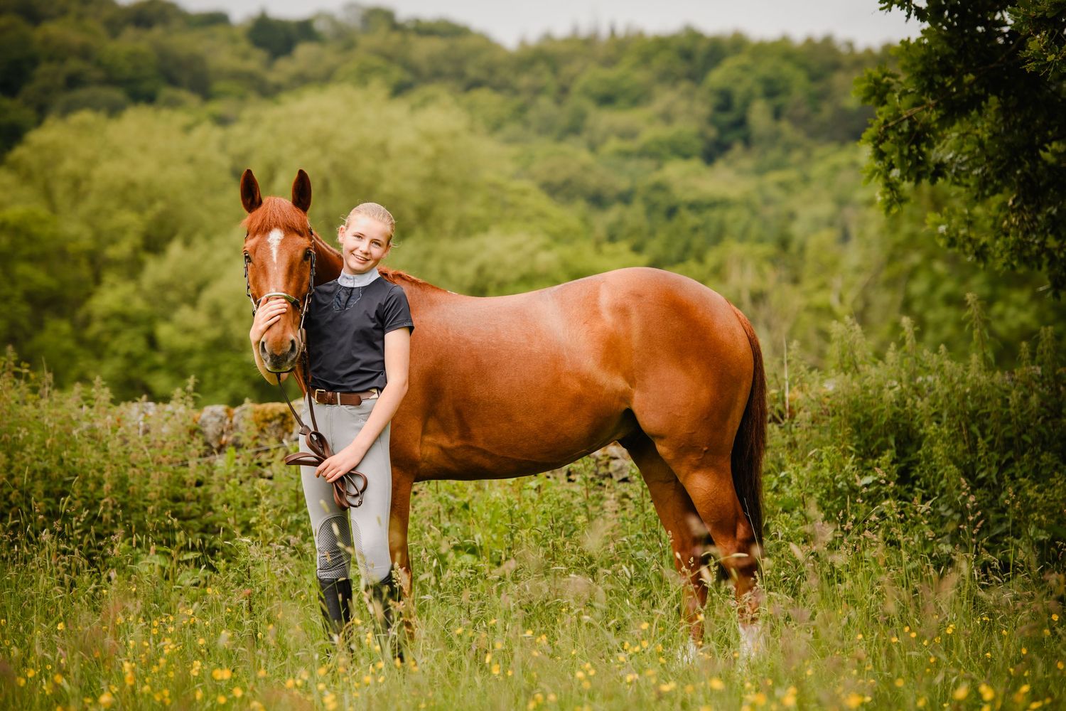 Young girl stands proudly beside a chestnut horse in a lush green meadow with wildflowers and trees in background.