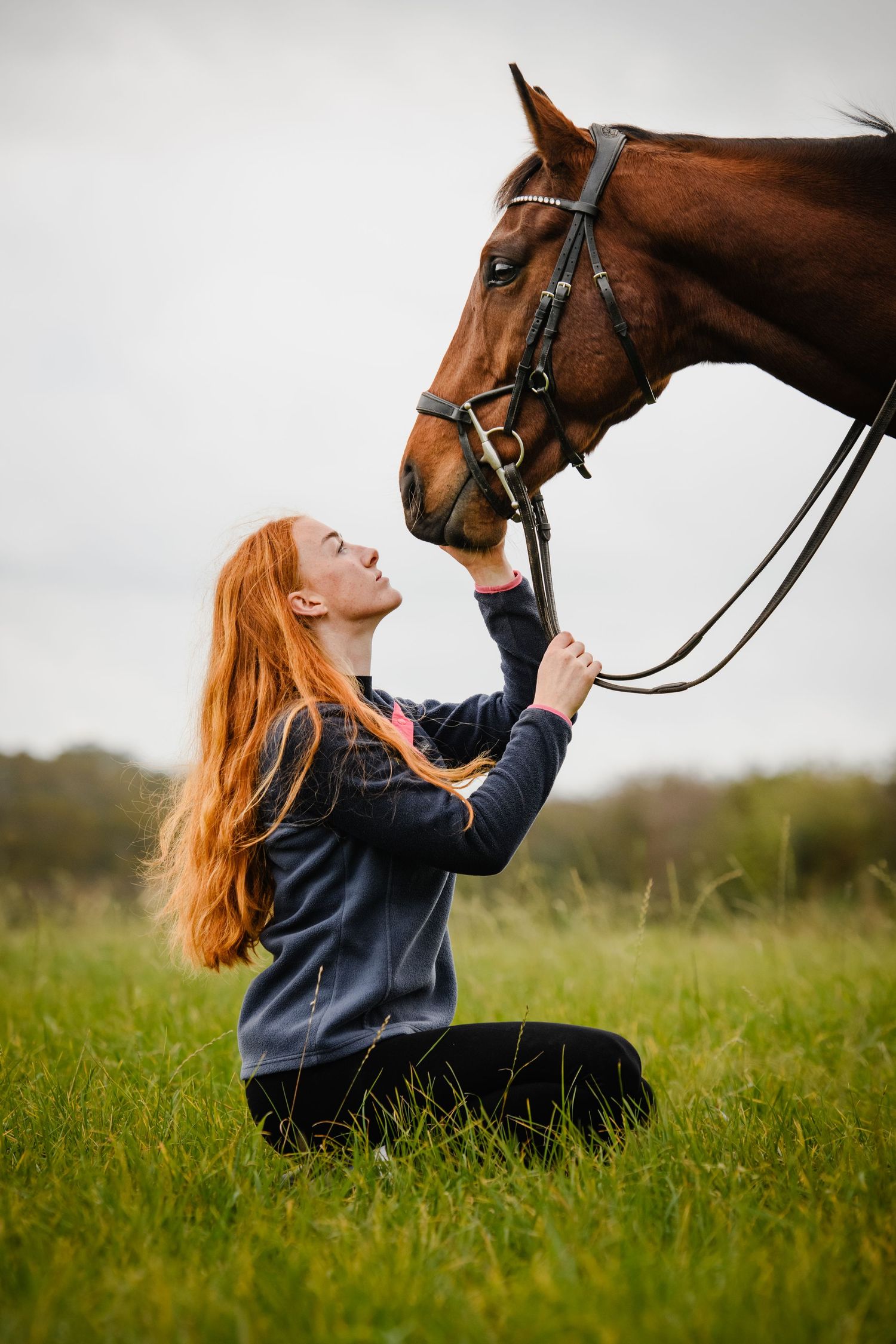A red-haired woman kneels in green grass, gently lifting a bay horse's nose toward her face.