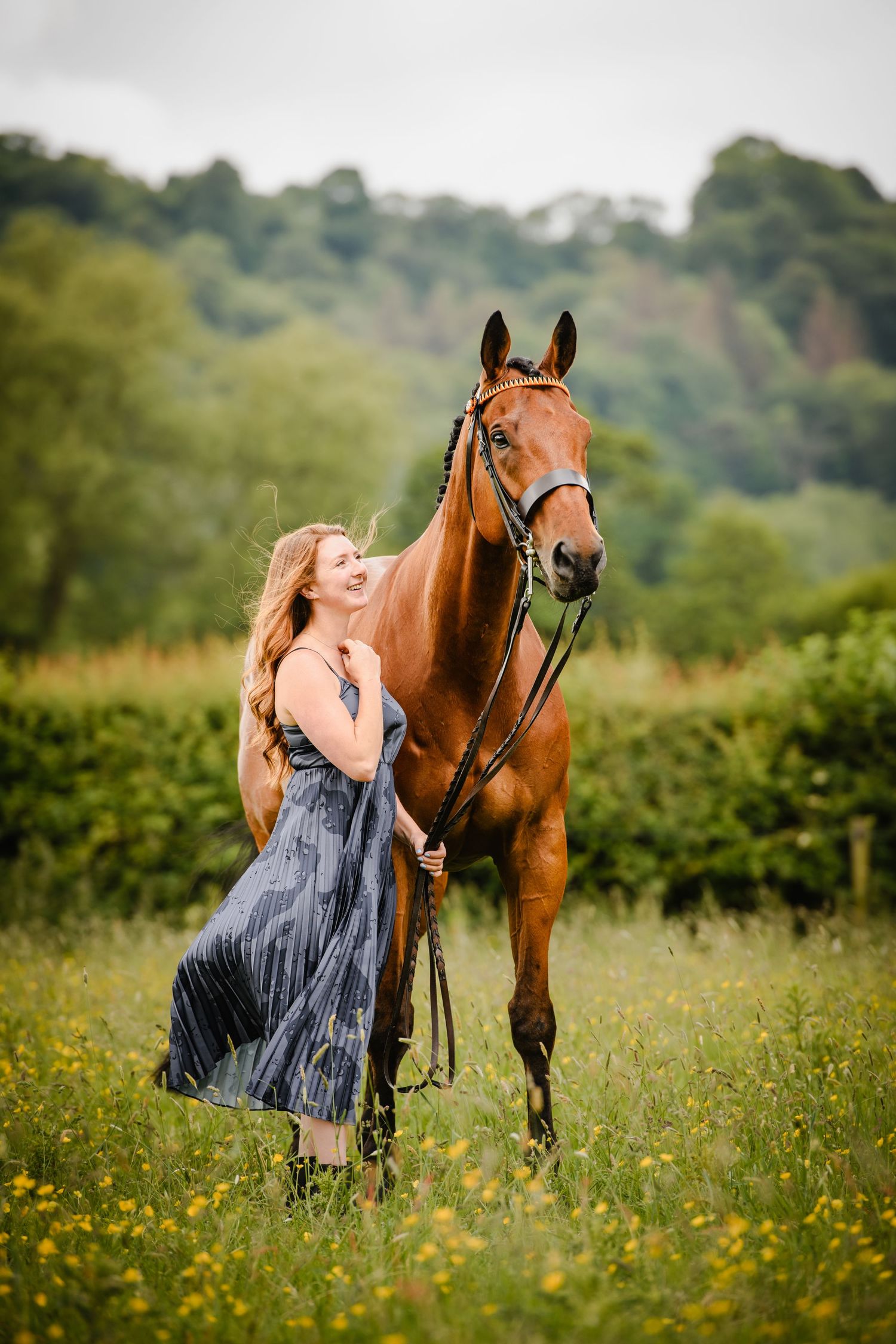 A smiling woman in a blue dress stands with a chestnut horse in a lush green field with yellow wildflowers.
