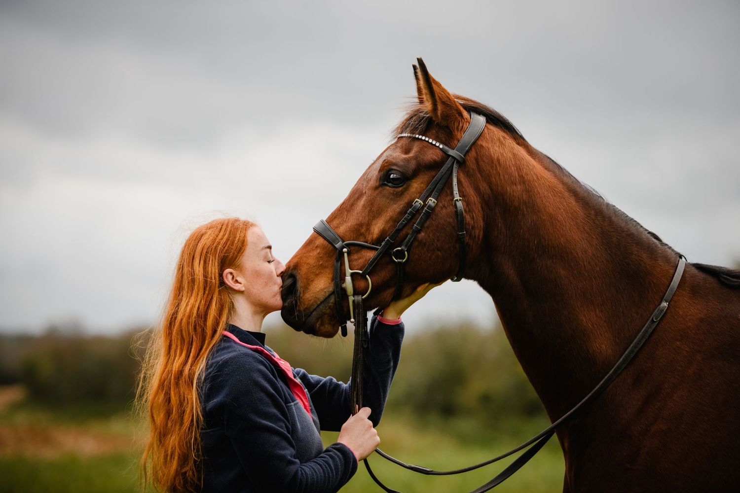 Red-haired woman sharing a tender moment with a chestnut horse outdoors under a cloudy sky.