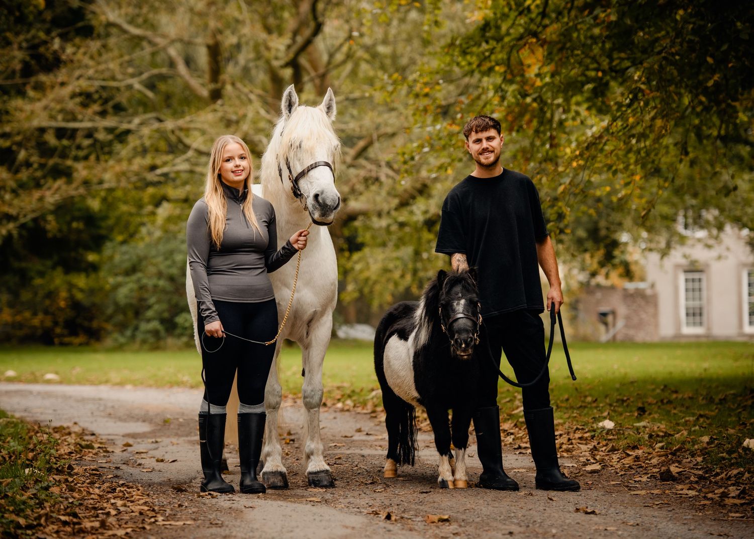 Two people standing with a white horse and black pony in an autumn park.