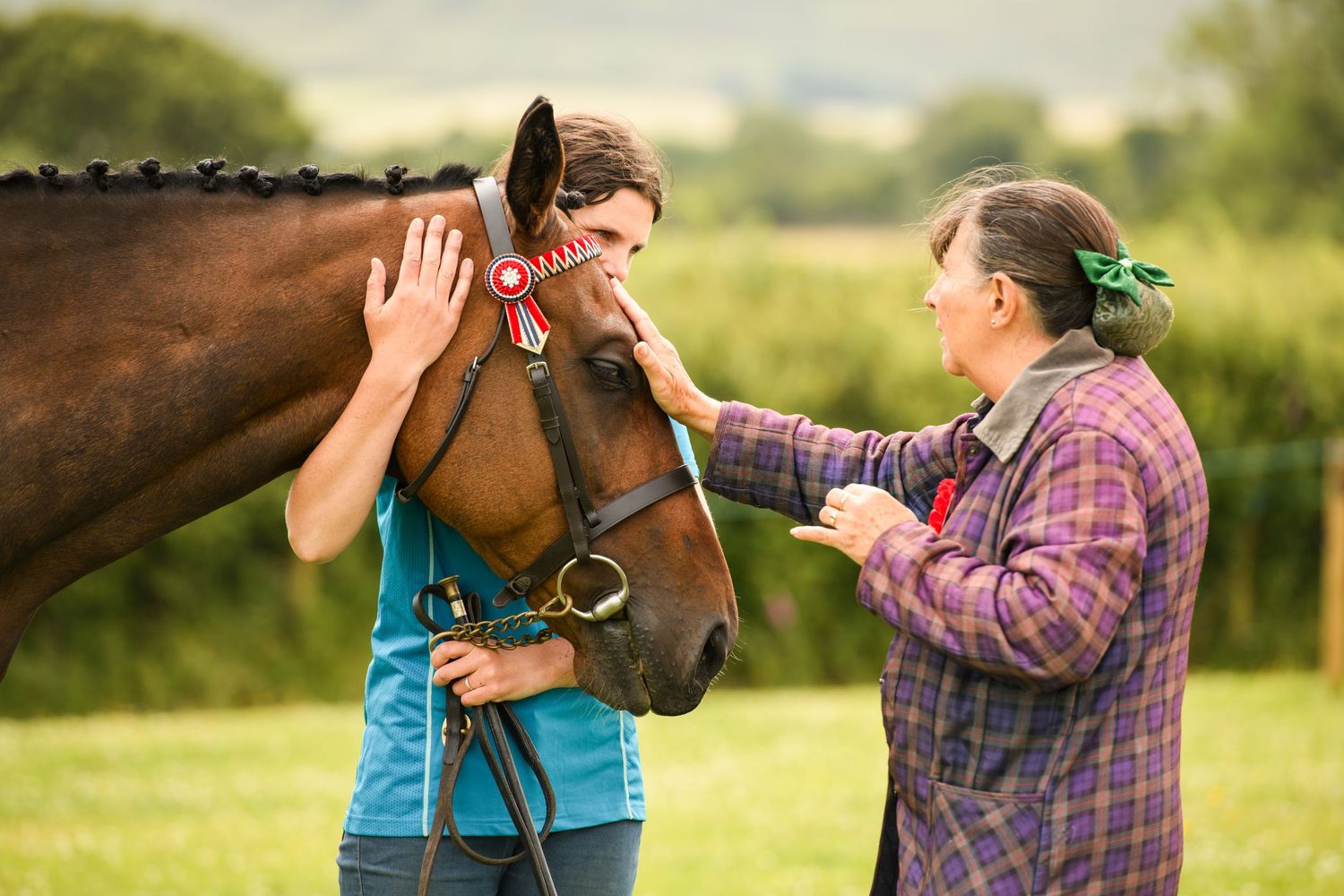 Two women proudly display a red rosette ribbon on a bay horse at an outdoor equestrian show.