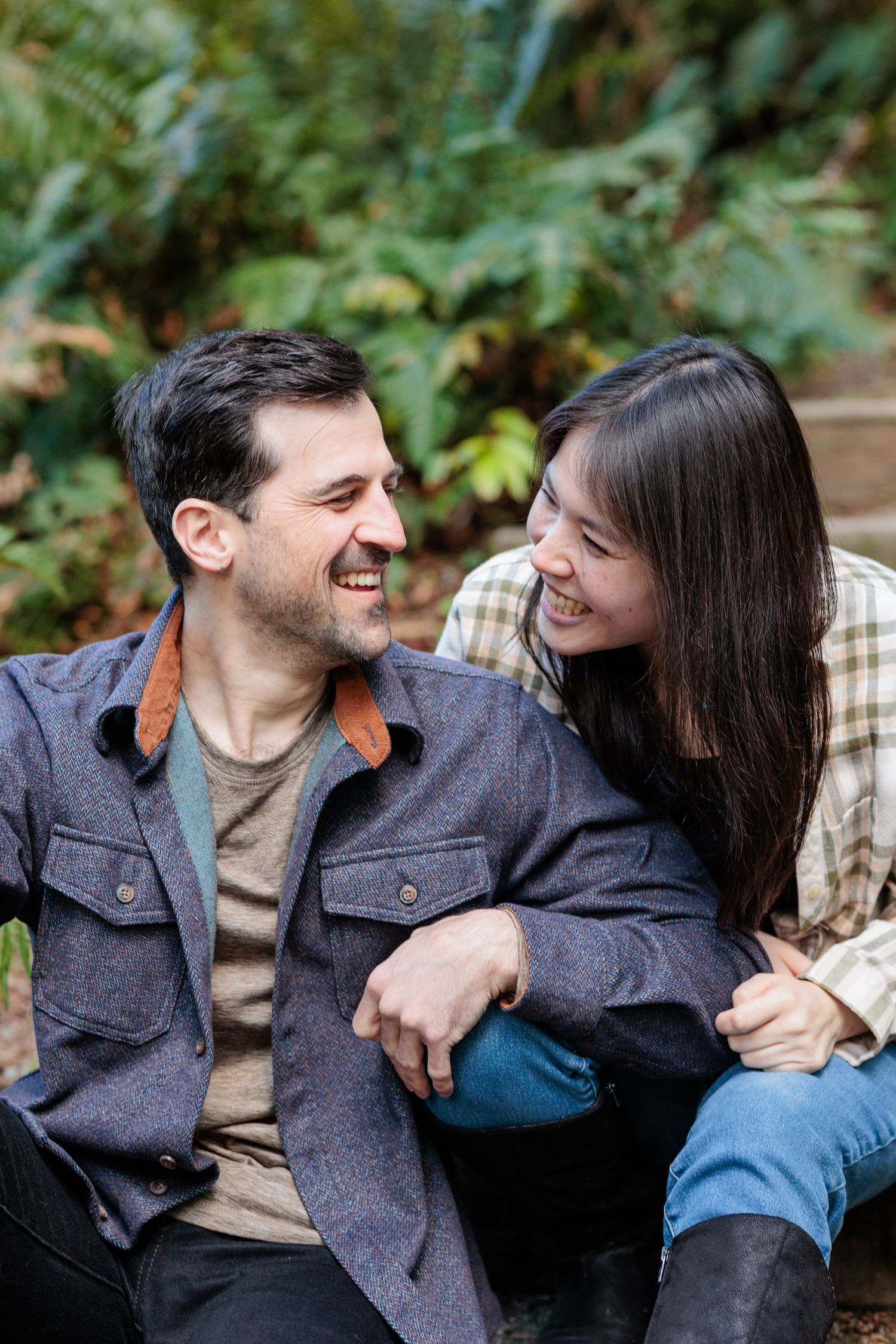 A smiling couple sitting outdoors in a lush green garden, laughing and looking at each other affectionately.