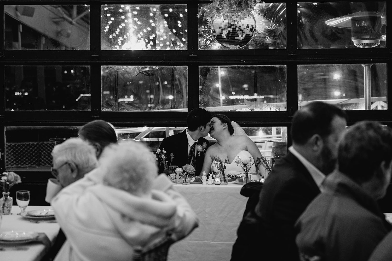 Newlyweds kissing at sweetheart table surrounded by wedding guests in black and white photo.
