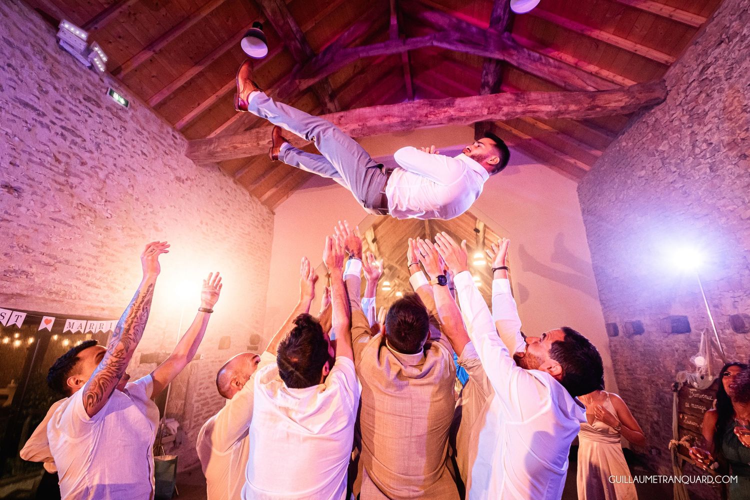 Party crowd dancing under purple lighting in rustic venue with wooden beams.