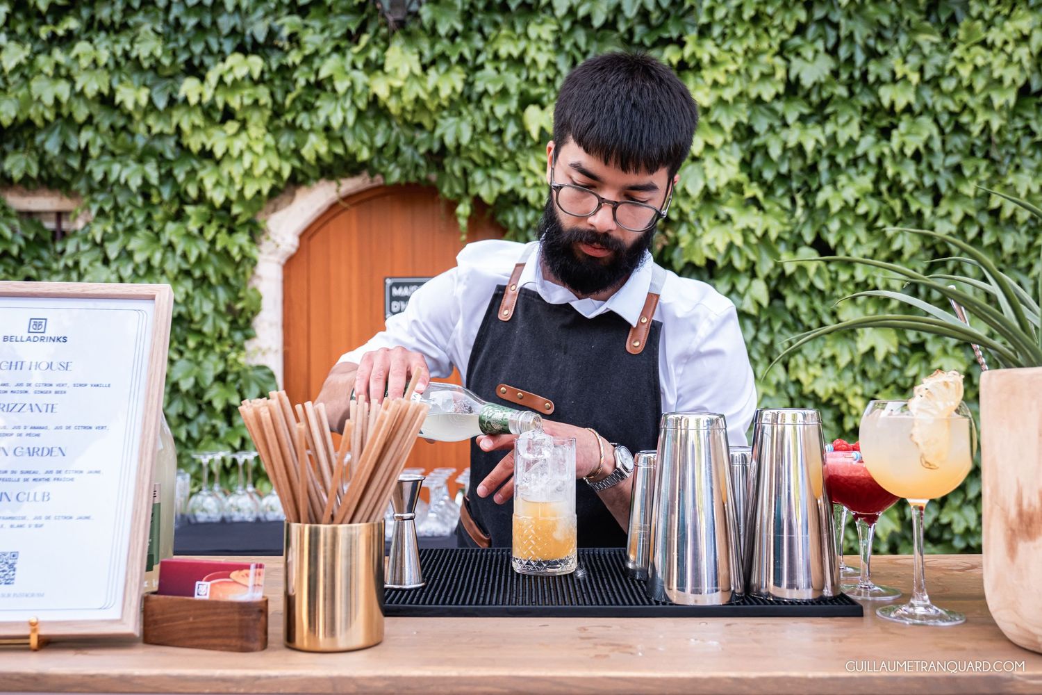 Bartender serving drinks at outdoor bar counter with green ivy wall backdrop.