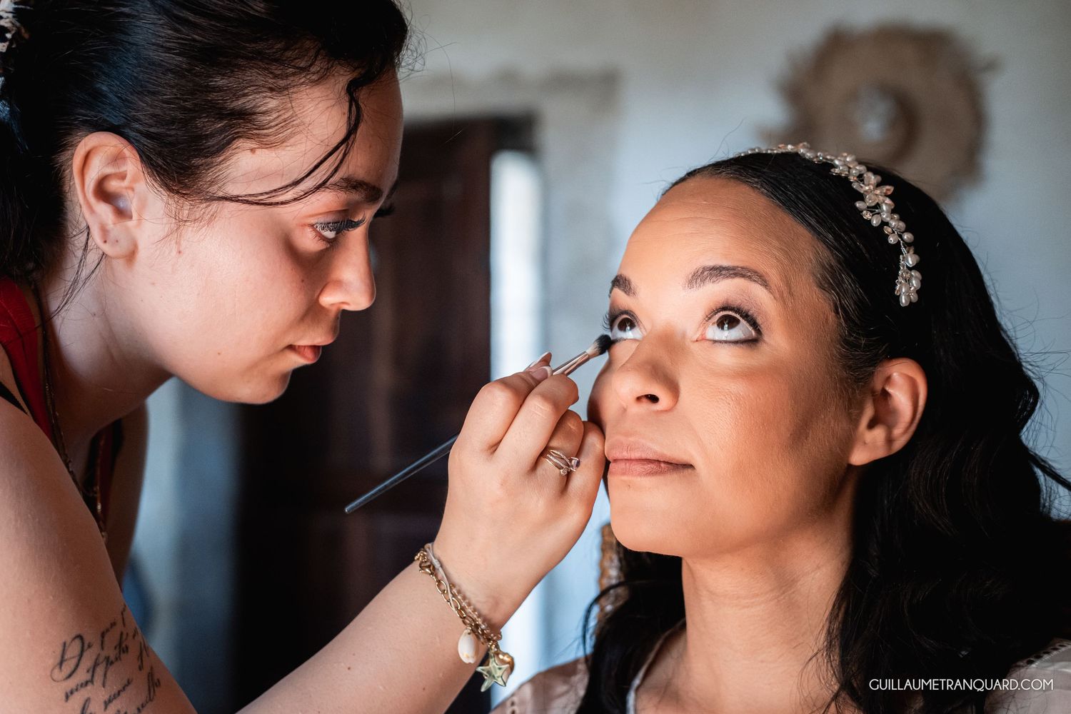 A makeup artist carefully applies eyeliner to a bride wearing a floral headband on her wedding day.