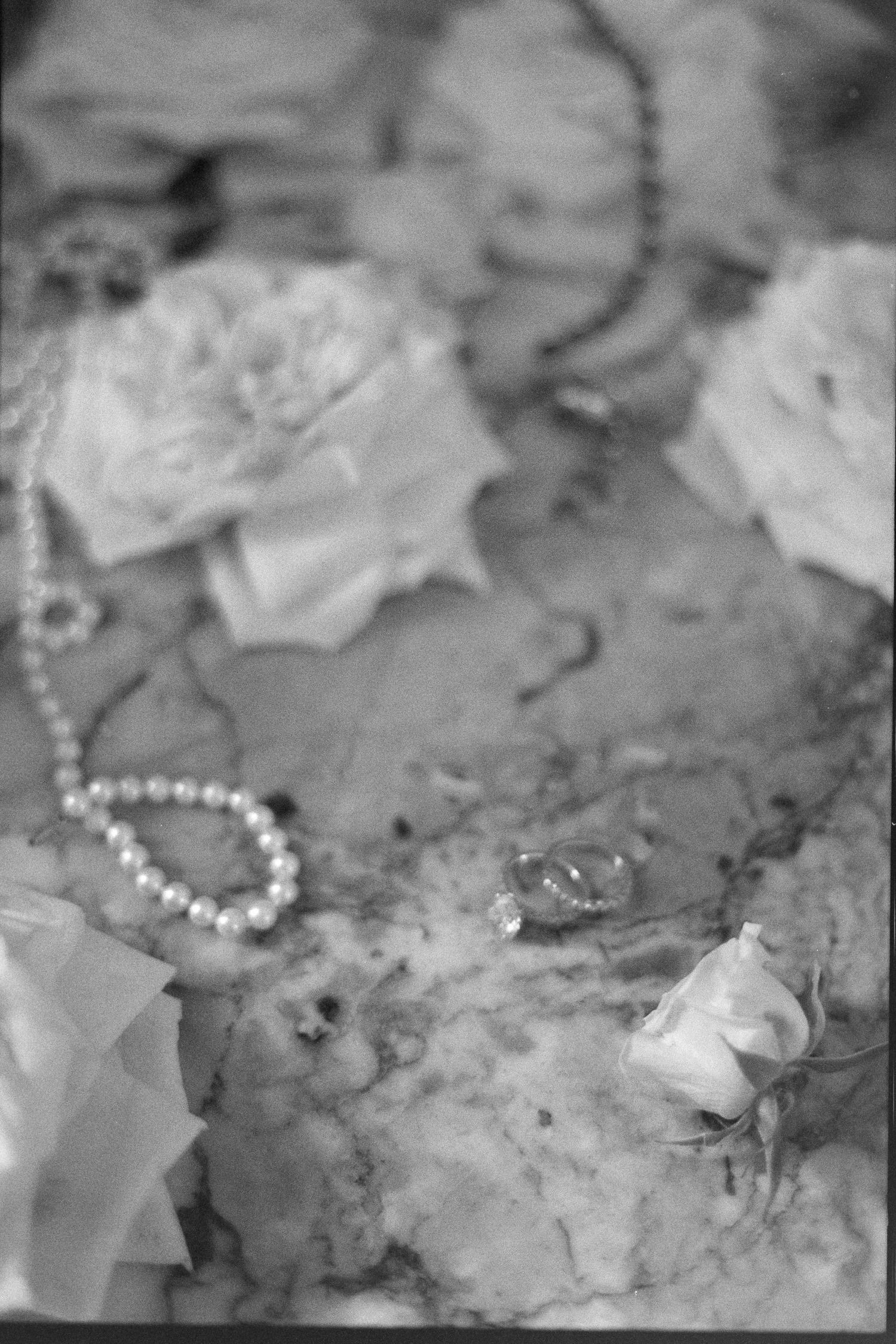 Black and white wedding details with scattered roses, pearl necklace, and rings on textured surface.