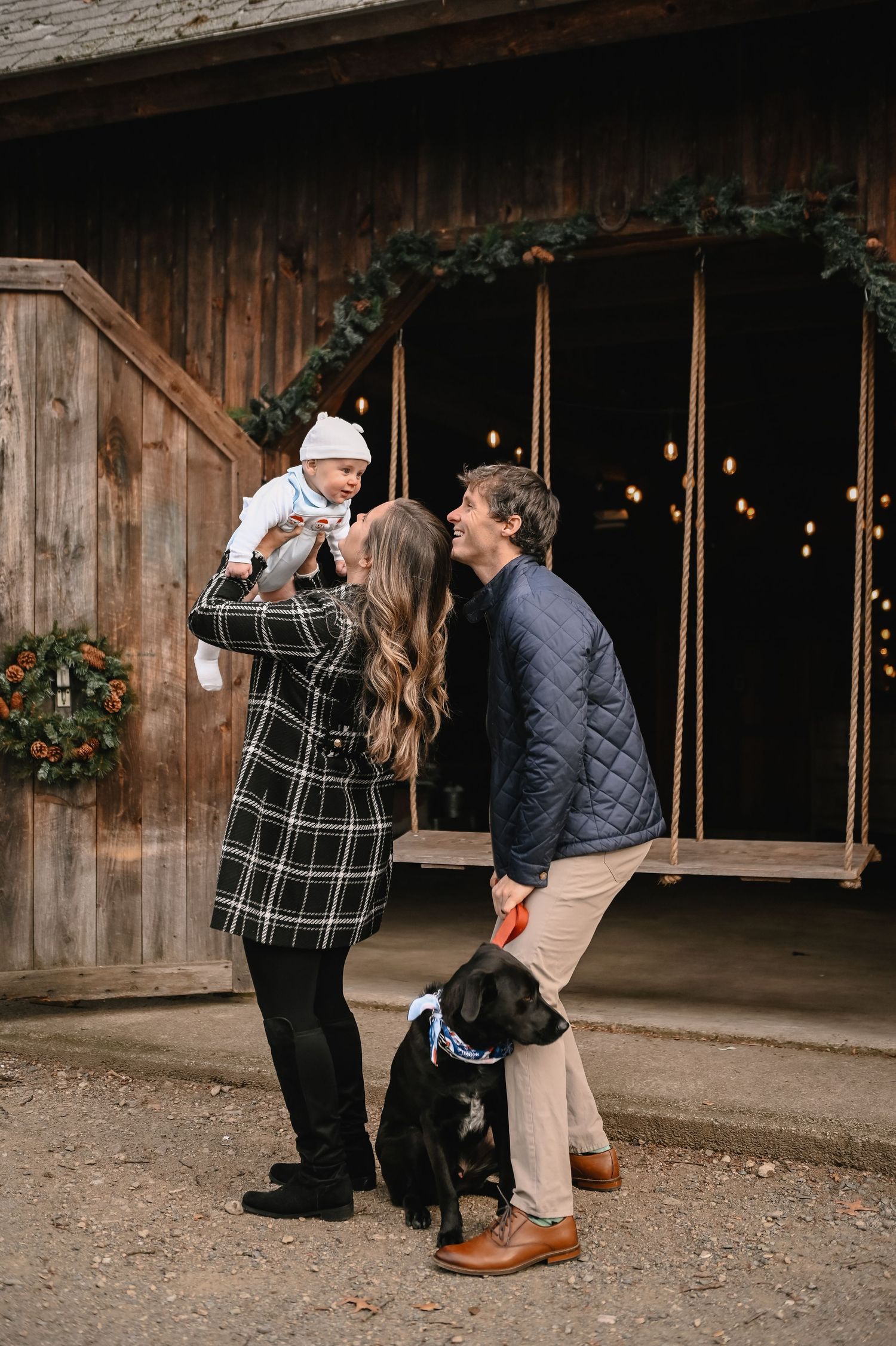 Family stands together outside rustic barn with their black dog on autumn day among string lights and greenery.