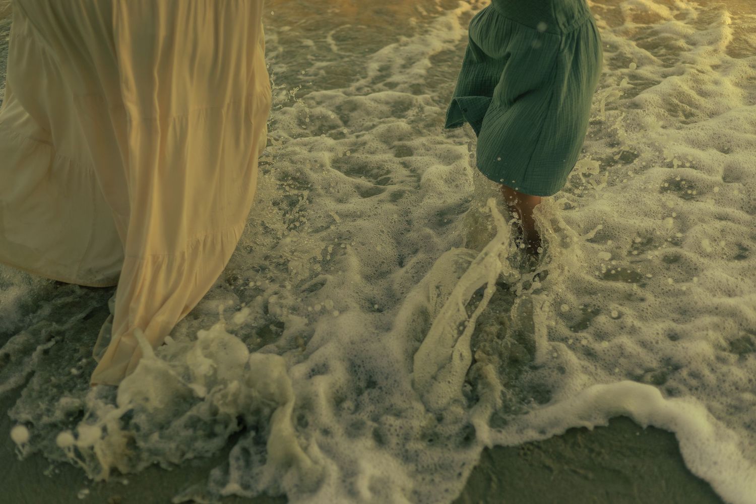 Ocean waves wash over legs and flowing fabric on a sandy beach during golden light during a storytelling family photography session.
