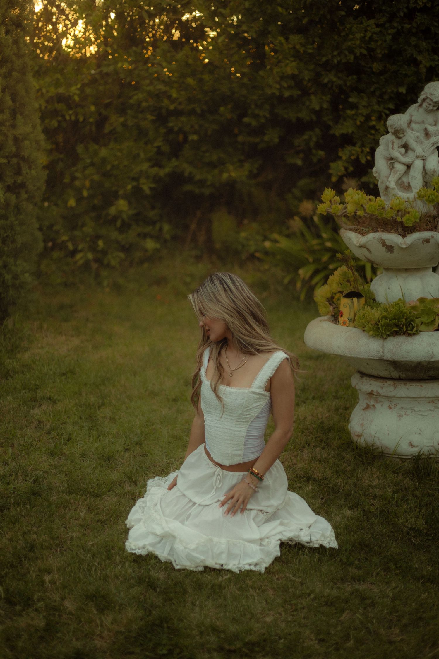 A woman in a white dress sits in a green dreamy garden during golden hour at a Victorian home in Los Angeles, California.