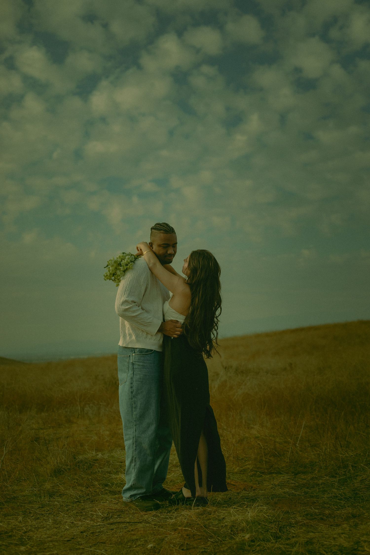 A romantic couple embraces on a hilltop against a dramatic cloudy sky at golden hour in Riverside California