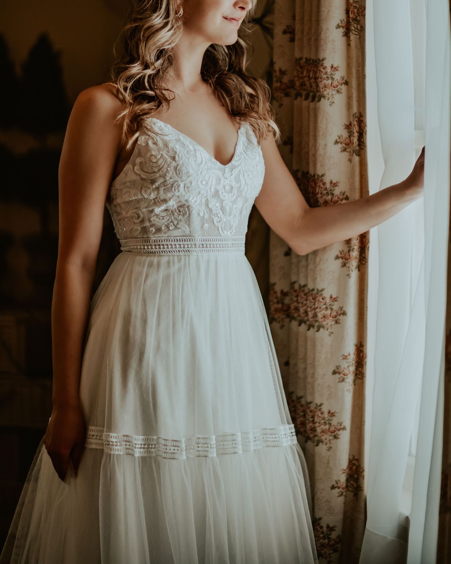 Bride standing next to the window in the bridal suite looking to see if she can find her groom as the sun shines on her wedding dress.