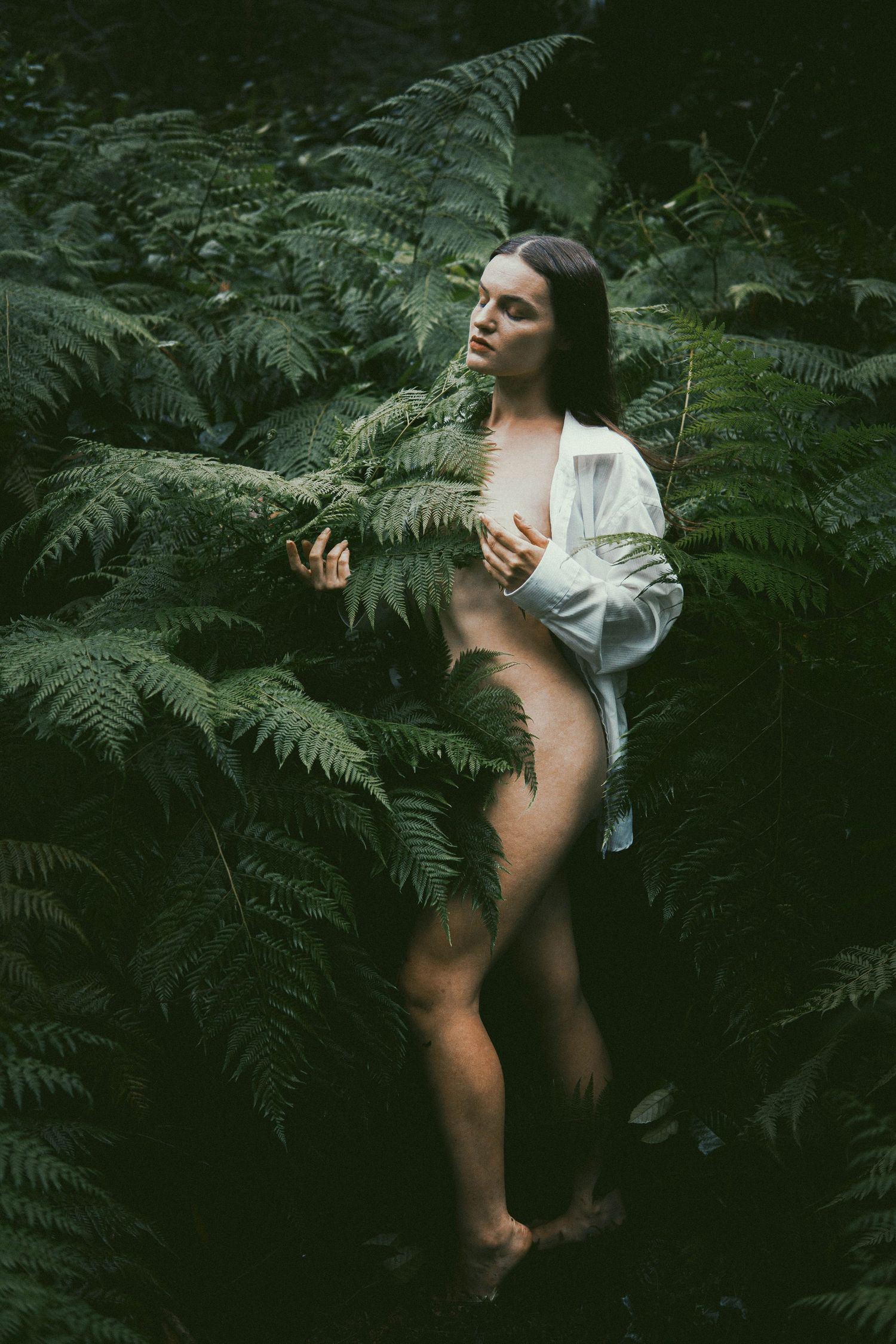 A woman in an open white shirt stands among lush green ferns in a dark forest setting.