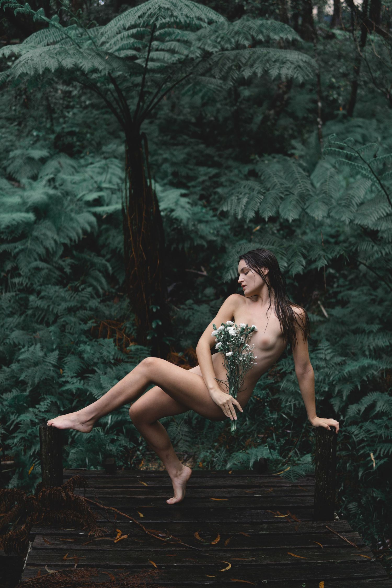 Nude woman sitting on wooden bench surrounded by lush green ferns and tropical foliage holding white flowers.