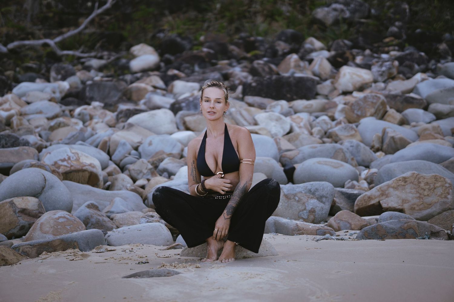 Woman in black bikini top and pants crouching on sandy beach with large rocks in background.