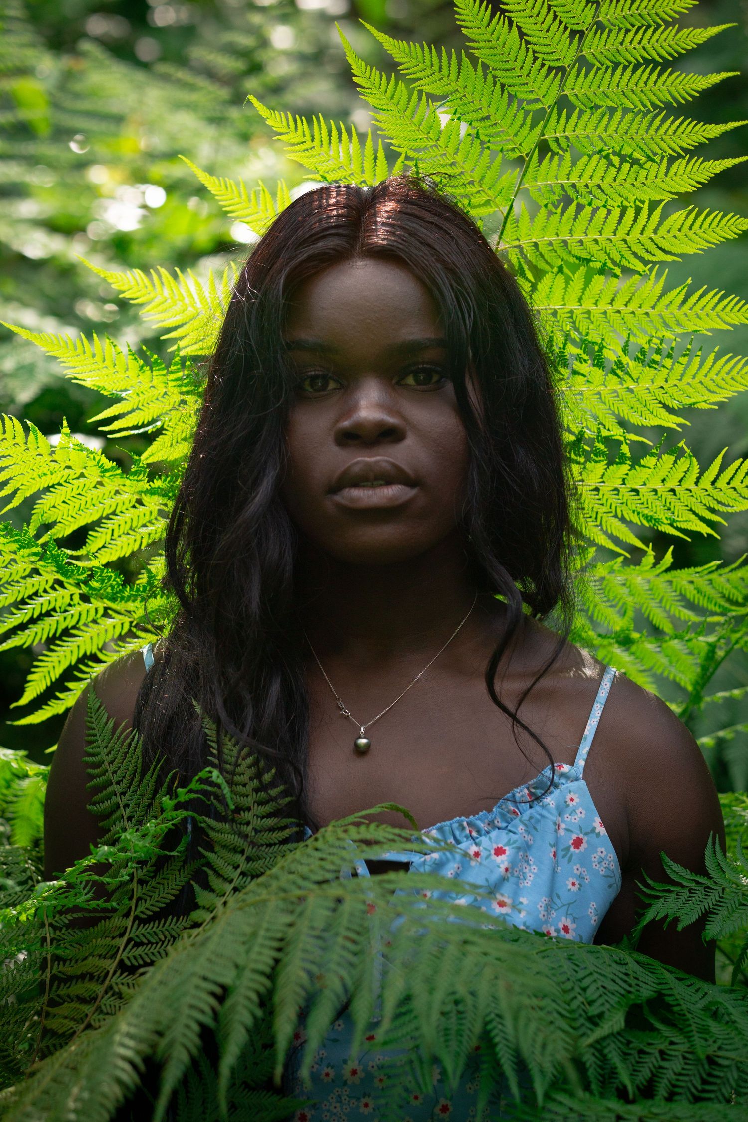 Young Black woman in blue floral dress surrounded by lush green ferns, looking directly at camera.