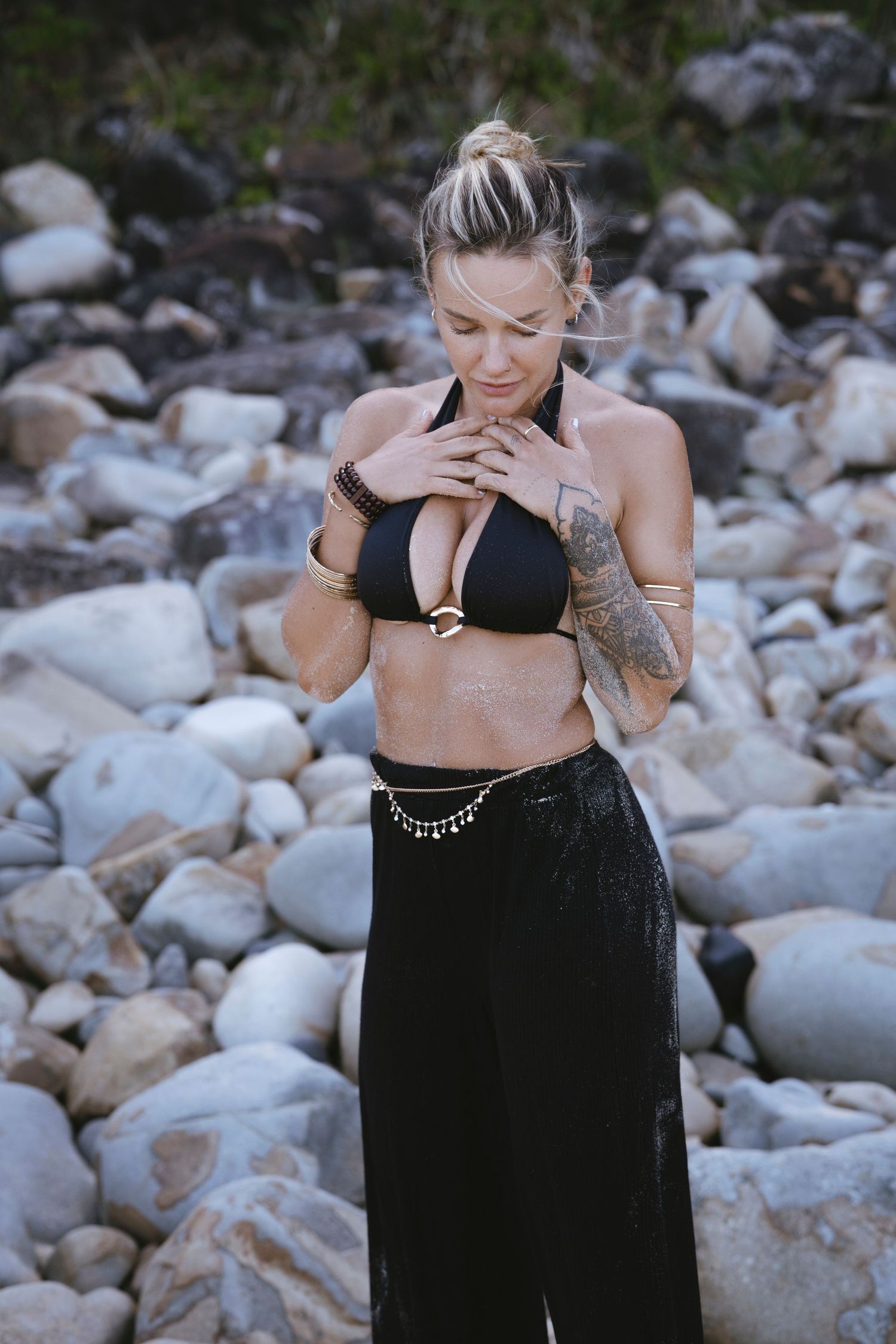 A tattooed blonde woman in a black bikini top and maxi skirt poses on a rocky beach.