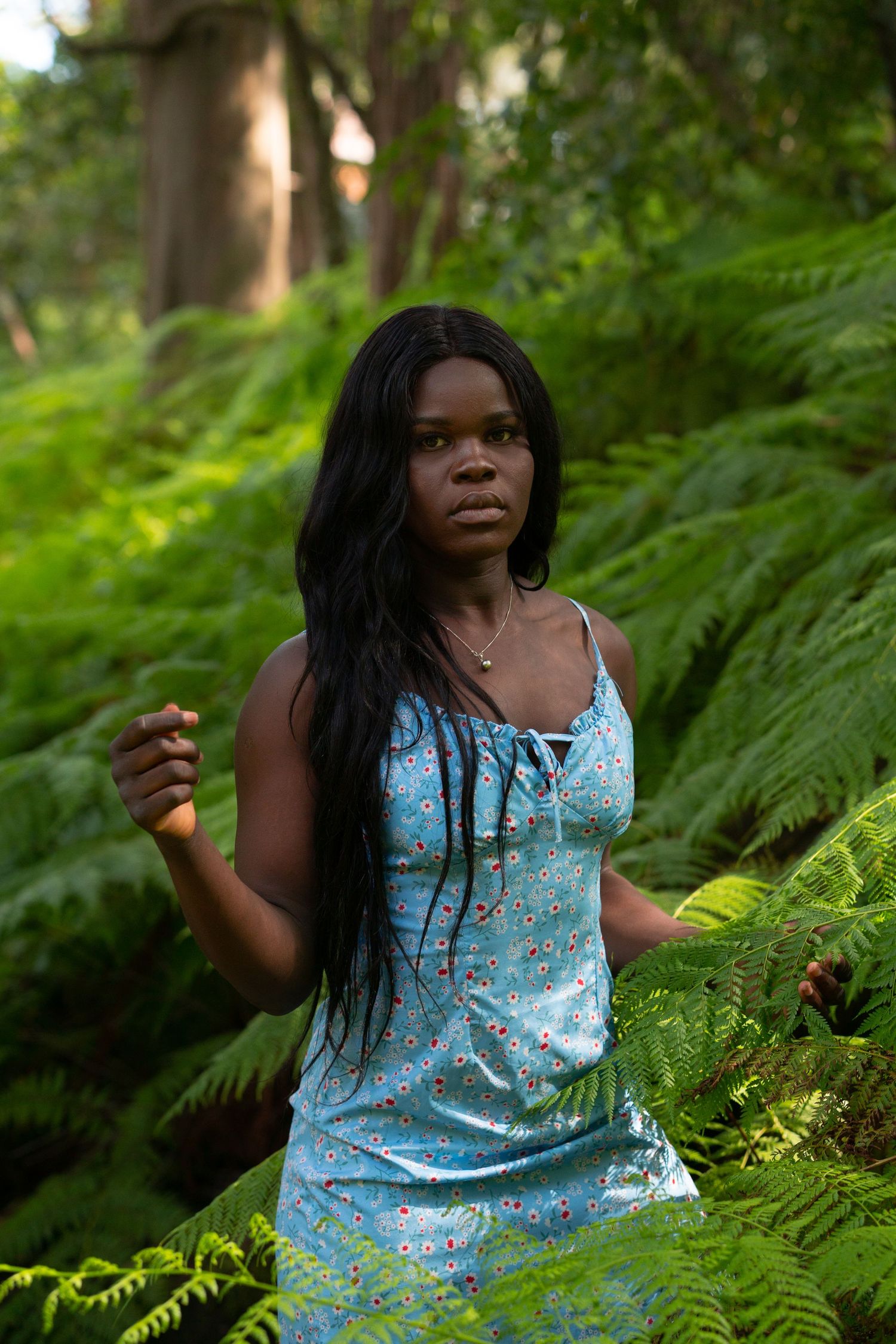 Young Black woman in blue floral dress standing among lush green ferns in a forest setting.