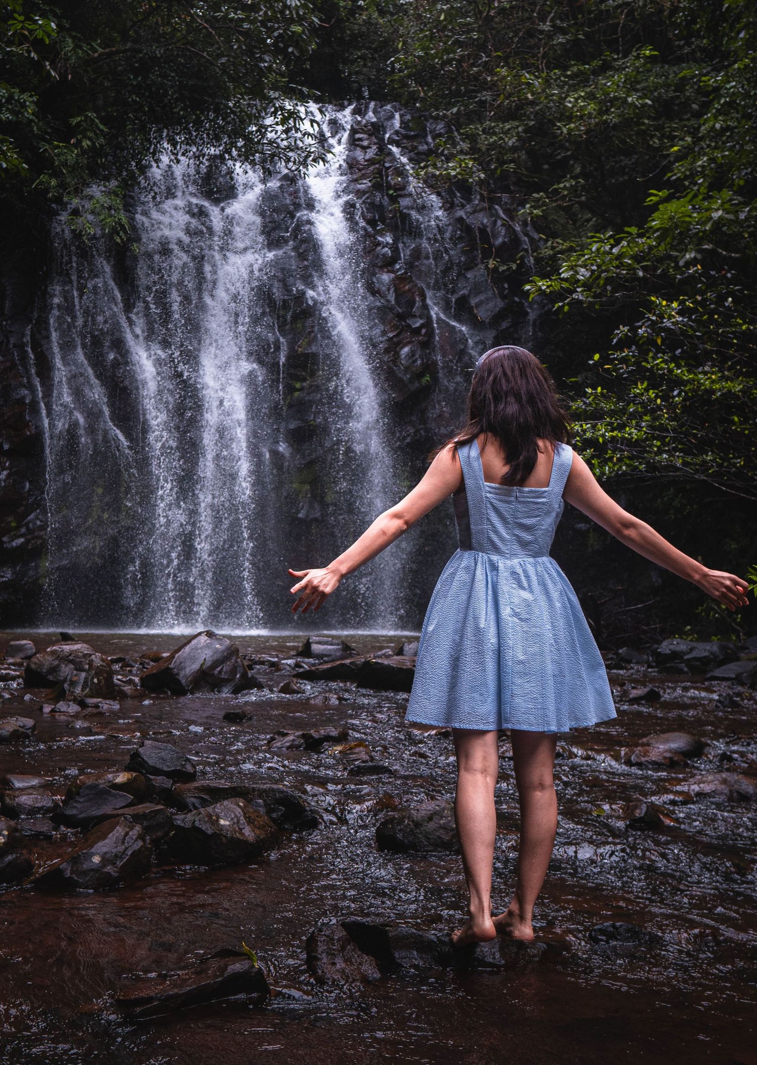 Woman in blue dress stands with arms outstretched before a lush waterfall surrounded by green trees and rocky terrain.
