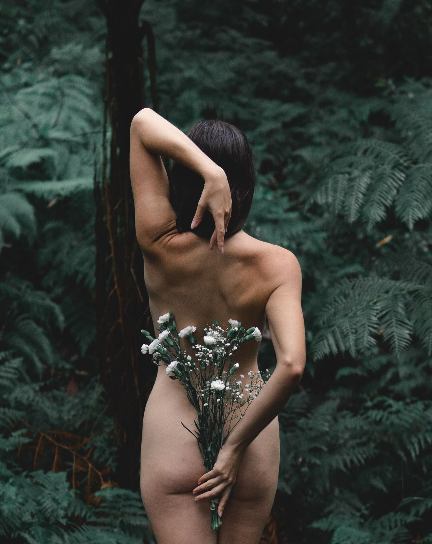 A nude woman holds white wildflowers in a lush green forest setting with ferns and trees.