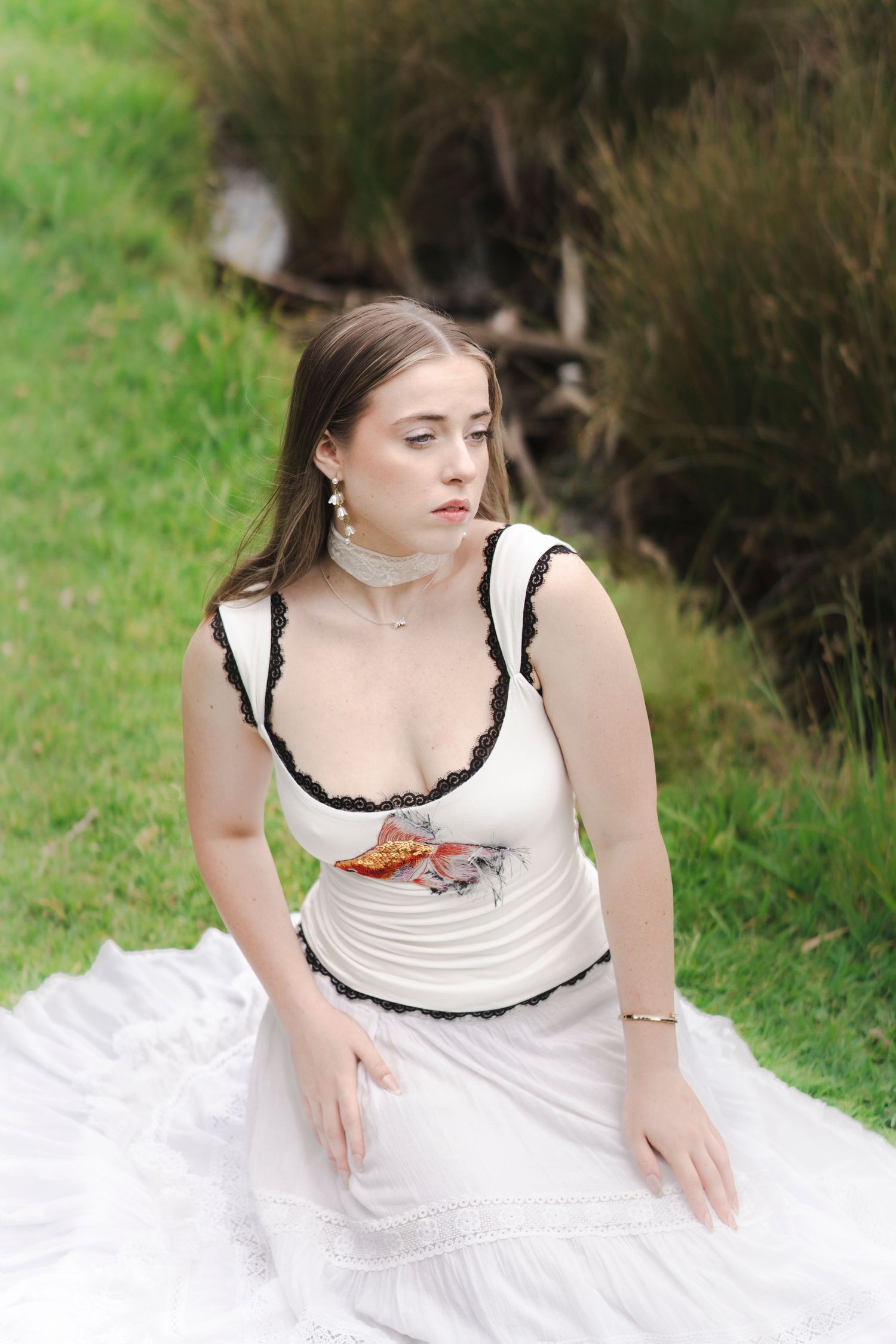 Young woman in white dress with black lace trim sits on green grass, wearing pearl earrings and bracelet.