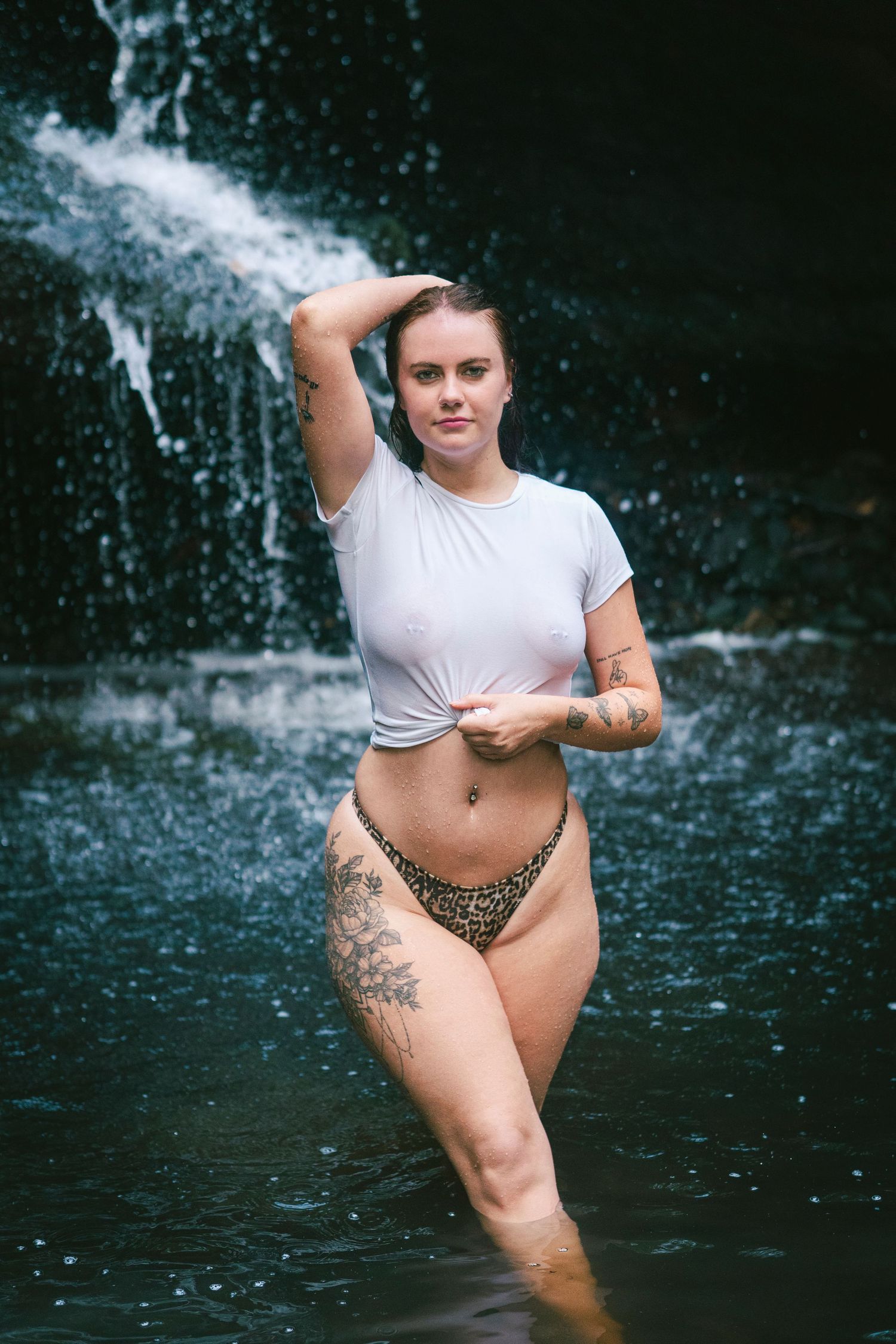 A tattooed woman in a white crop top and leopard bikini bottoms poses in a natural waterfall pool.