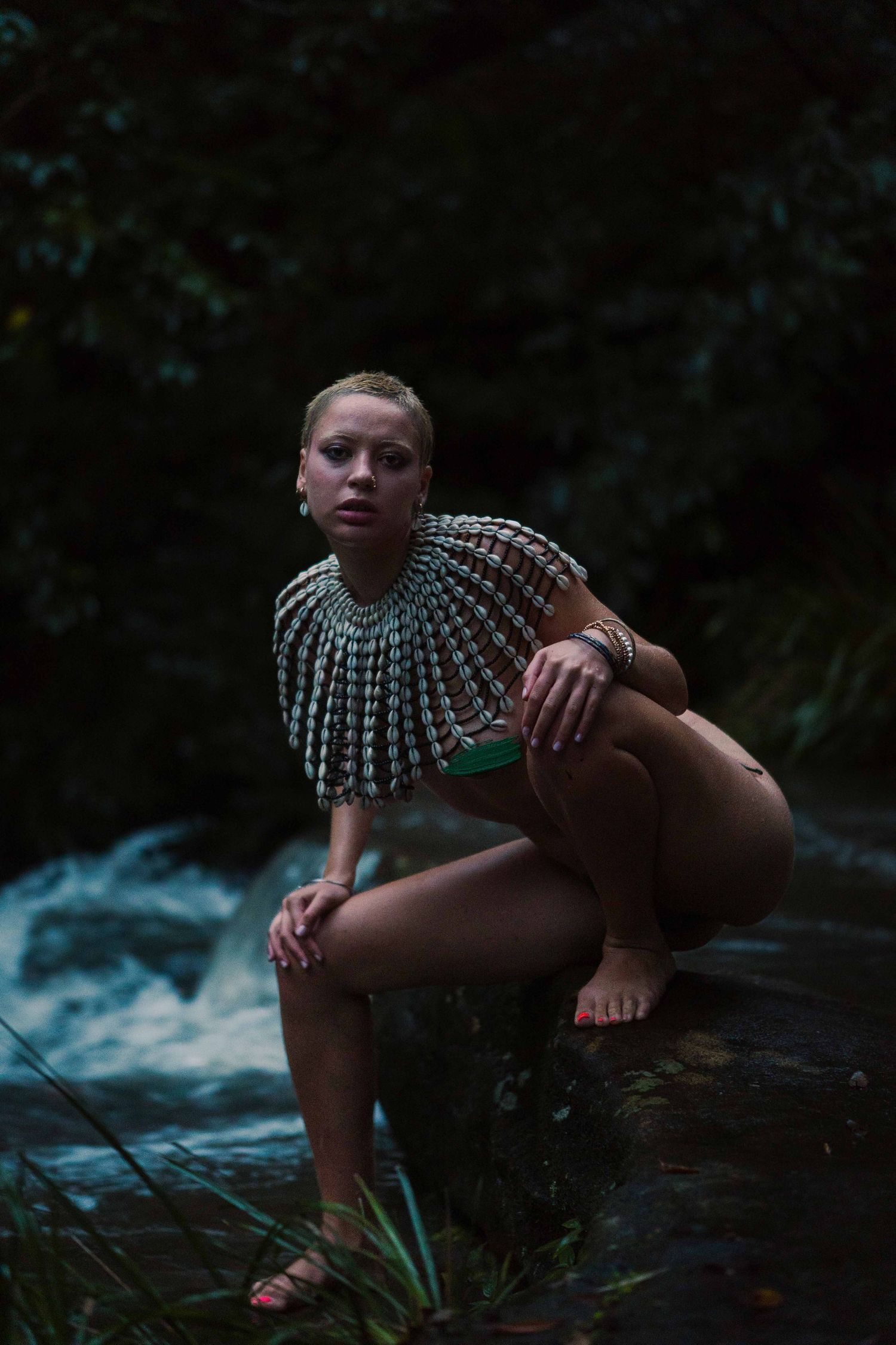 Woman crouching by a forest stream wearing an ornate silver chainmail cape and green bracelet.
