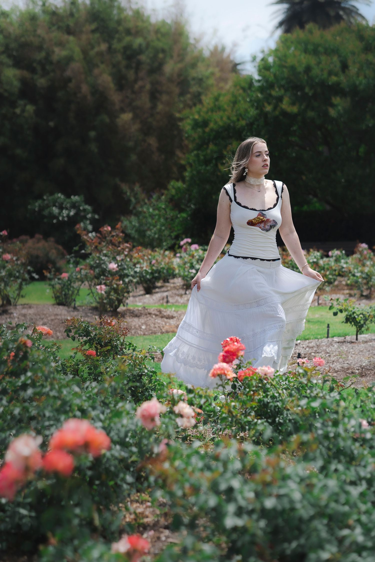 Woman in white maxi dress standing among colorful roses in a lush garden, gazing into the distance.