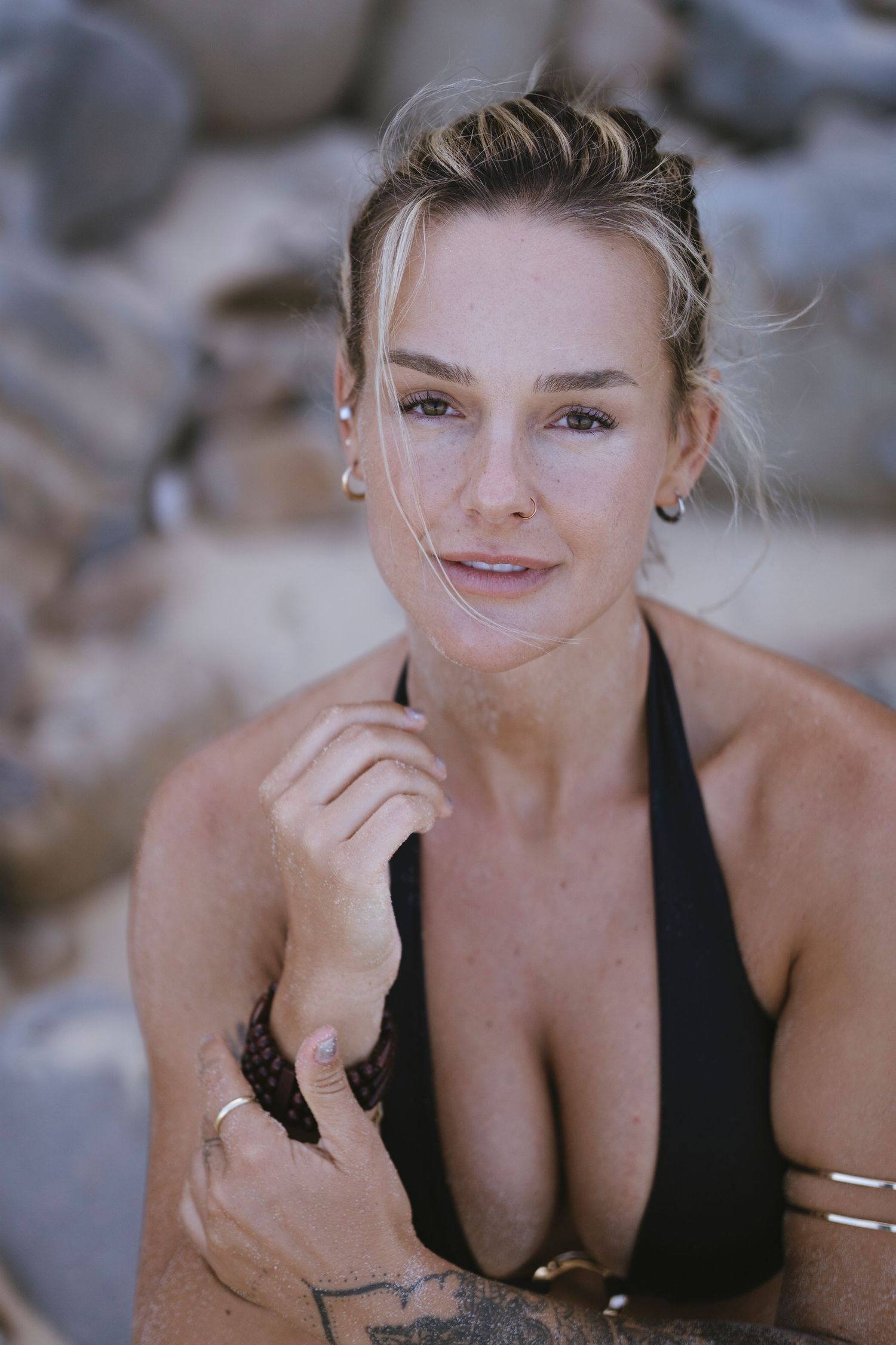 Woman in black bikini with tattoos posing near rocks on a beach, with natural makeup and hair up.