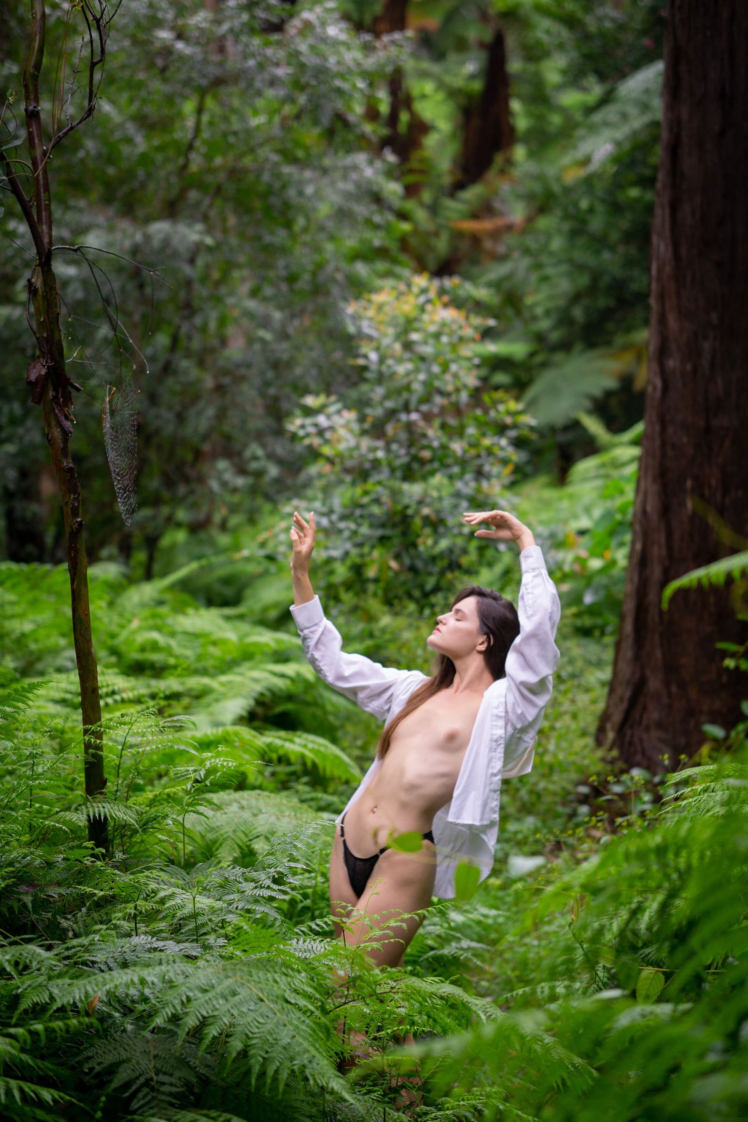 A woman in an open white shirt poses gracefully among lush green ferns in a dense forest.