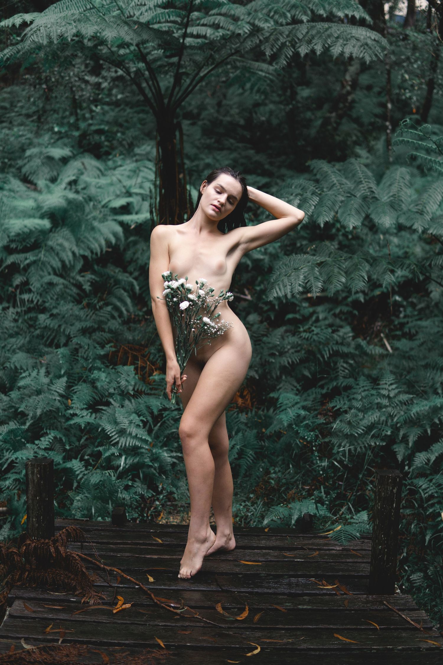A nude woman poses gracefully on a wooden deck surrounded by lush green ferns in a forest setting.