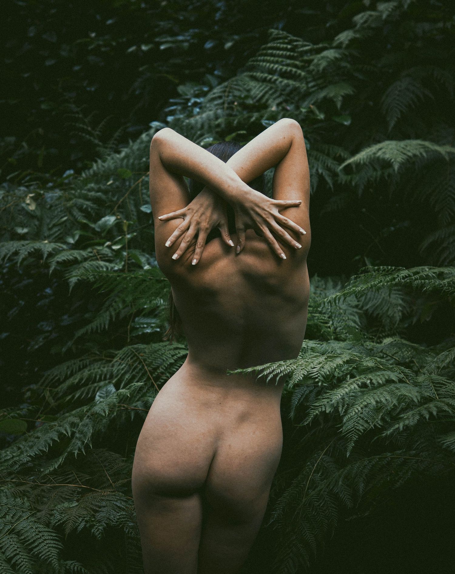 A nude figure stands with back turned among lush green ferns, hands crossed behind their head in nature.
