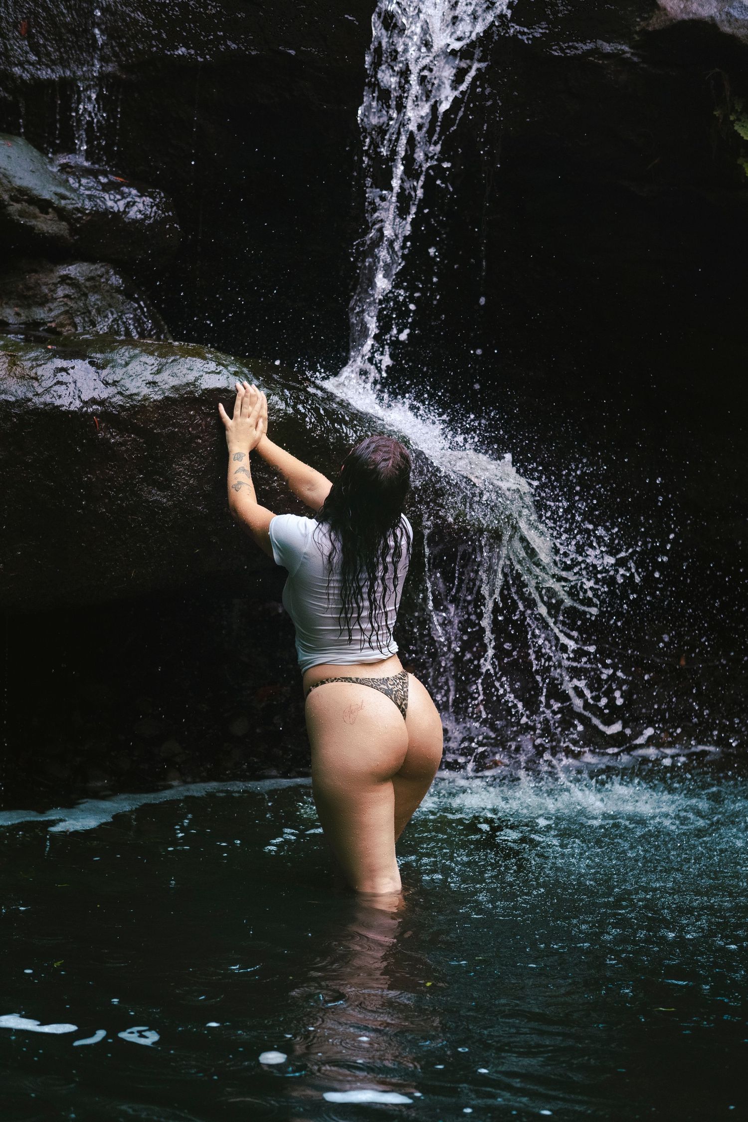 Woman in white top and bikini bottom standing in a natural pool beneath a cascading waterfall.