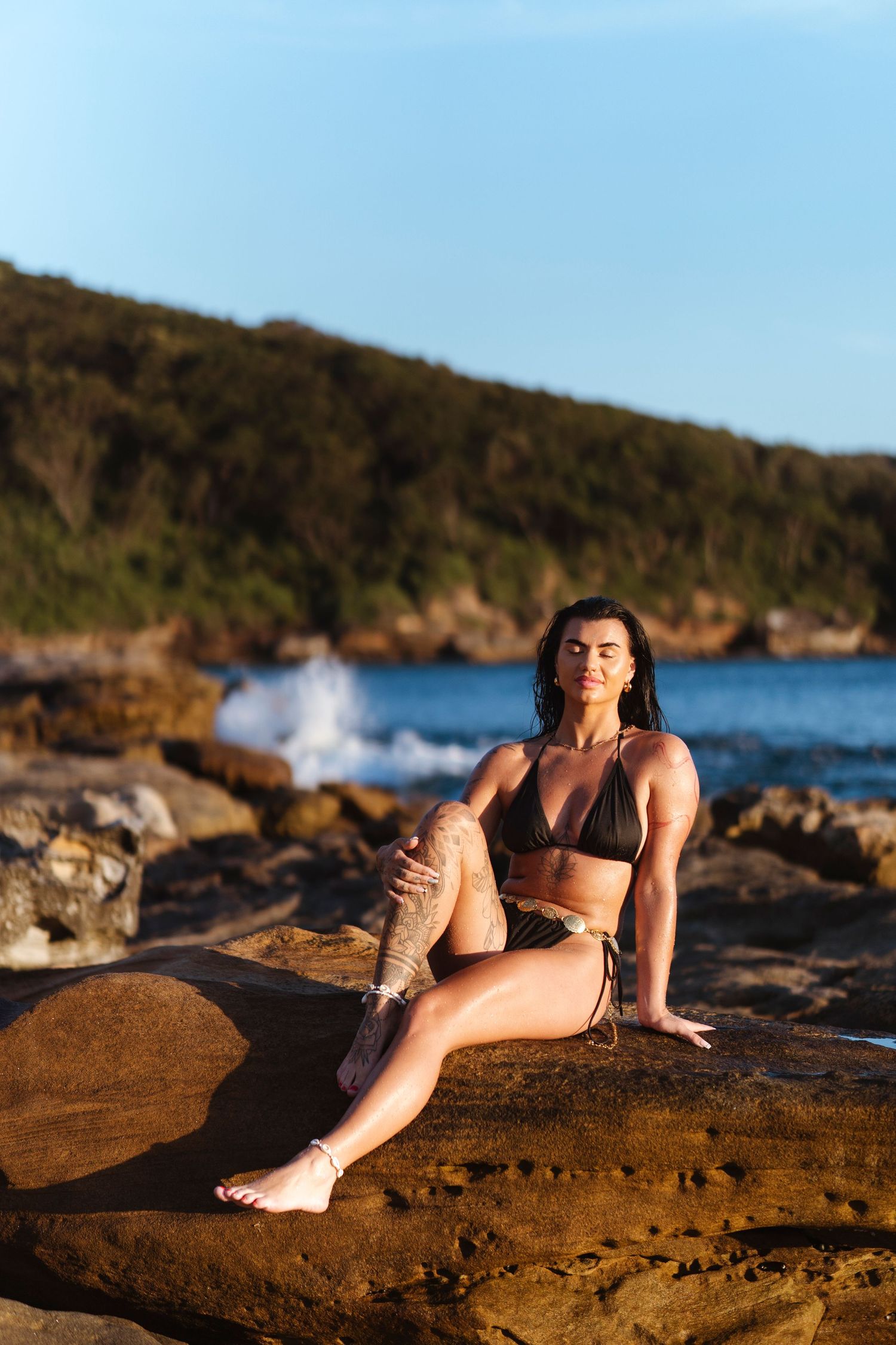 Woman in dark bikini poses on rocky shoreline with ocean waves and lush green hills in background.