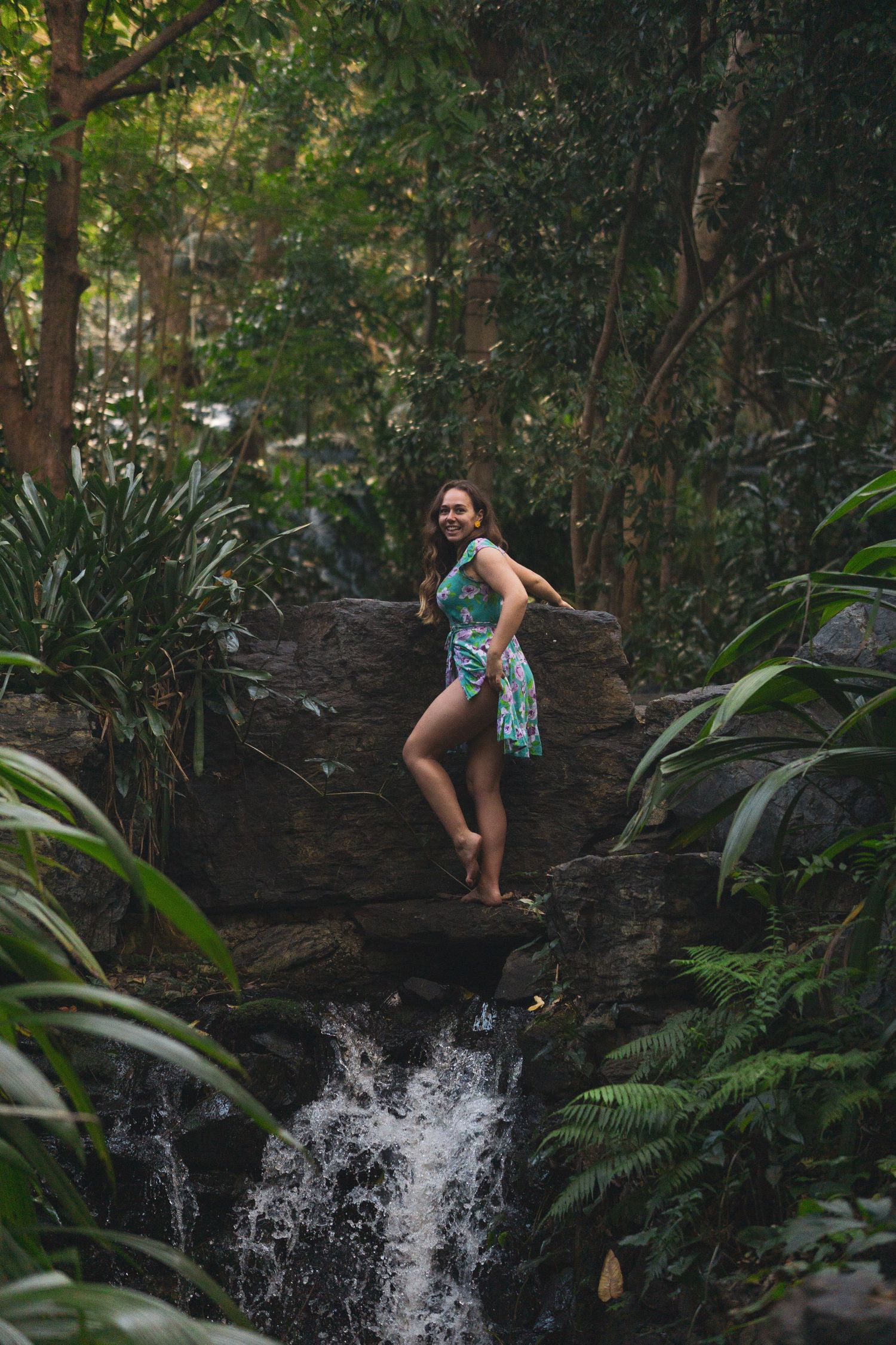 Woman in floral dress standing on rocks above a cascading waterfall surrounded by lush tropical forest greenery.