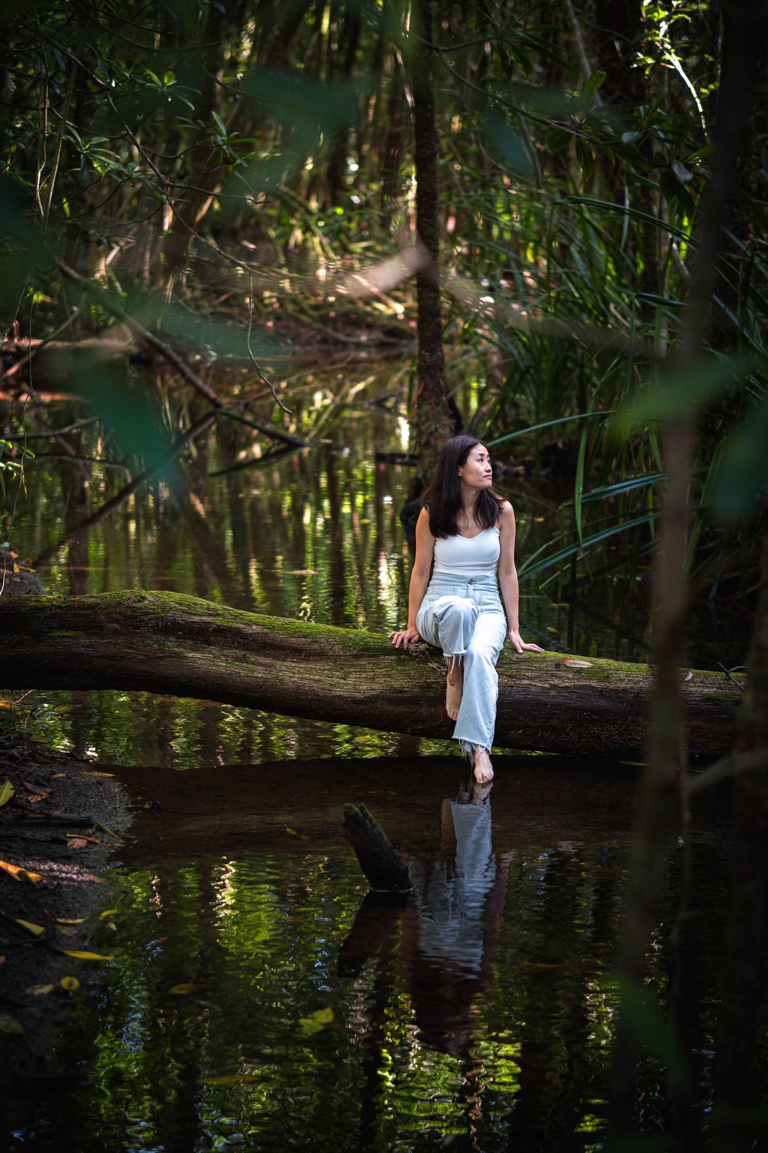 A young woman in white sits on a mossy log over a dark forest creek, her reflection visible in the still water below.
