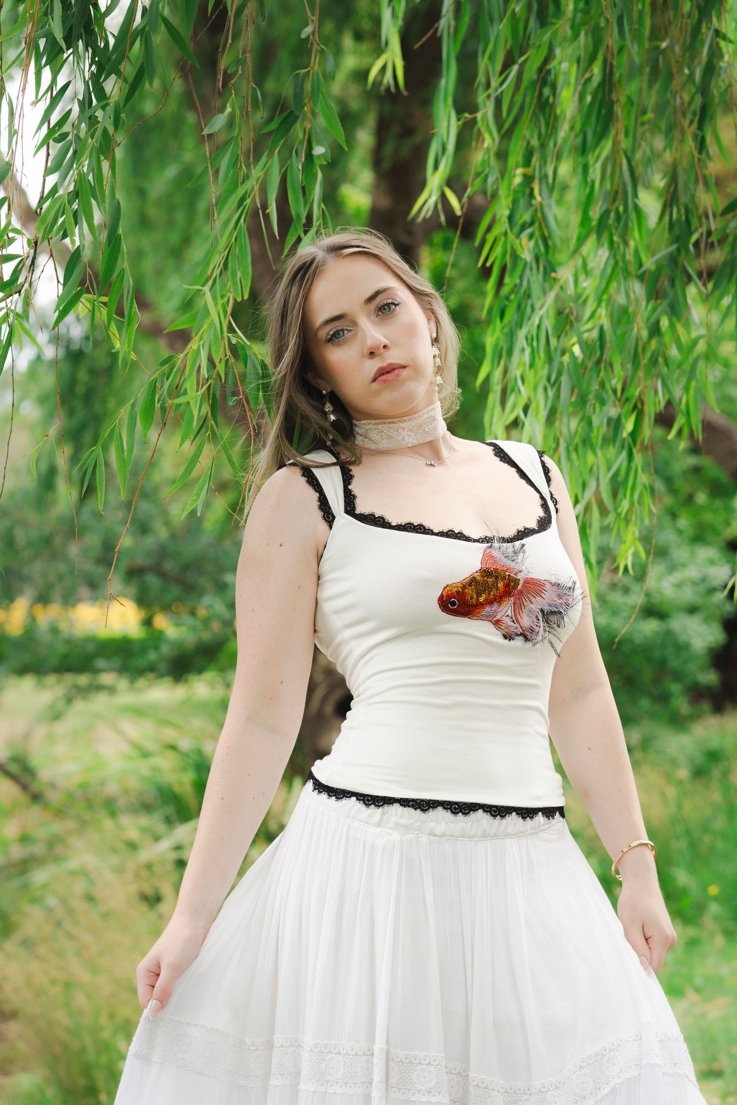 Young woman in white top and skirt holding a goldfish brooch, standing under a willow tree outdoors.