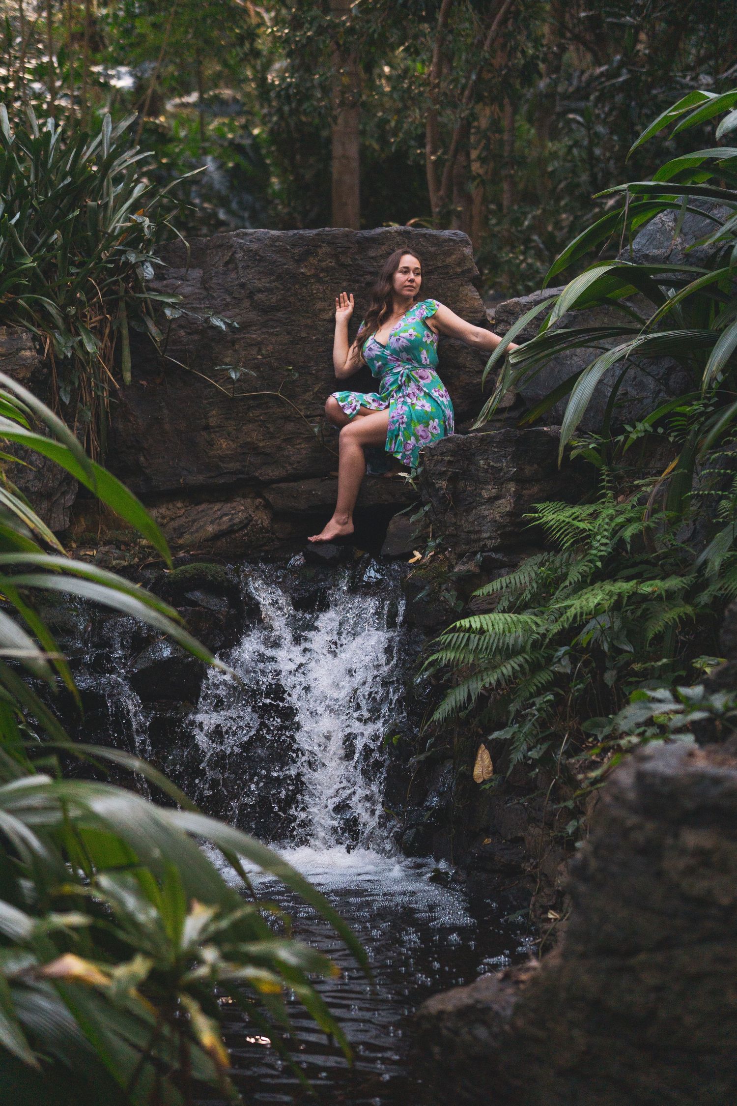Woman in floral dress sitting on rocks beside a lush tropical waterfall surrounded by green ferns and foliage.