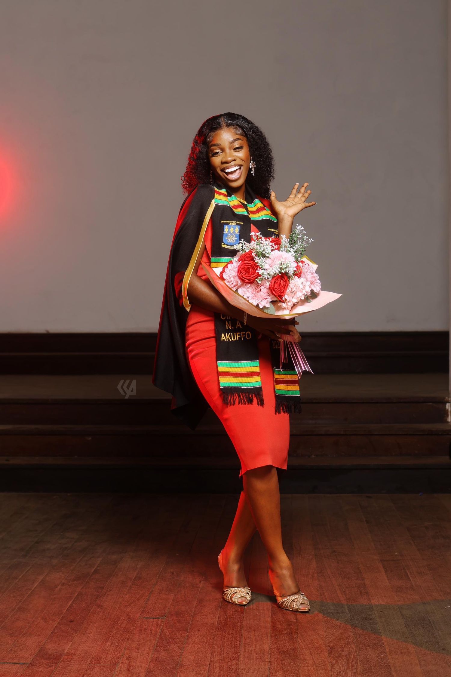 Graduate in red dress celebrating with flowers and stole on wooden stage.