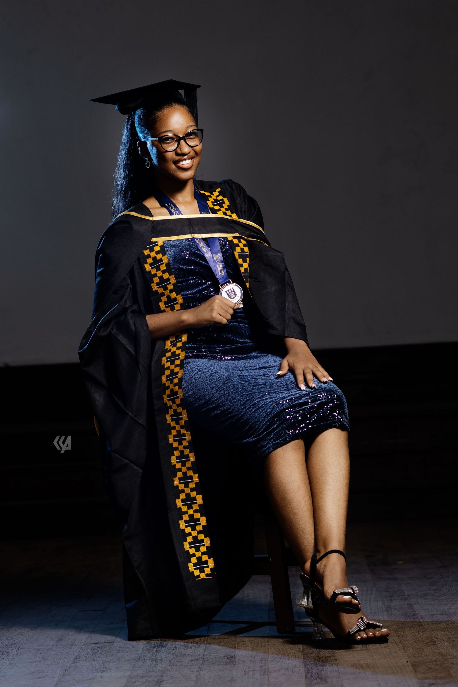 Graduate in navy dress and academic regalia posing against dark backdrop.