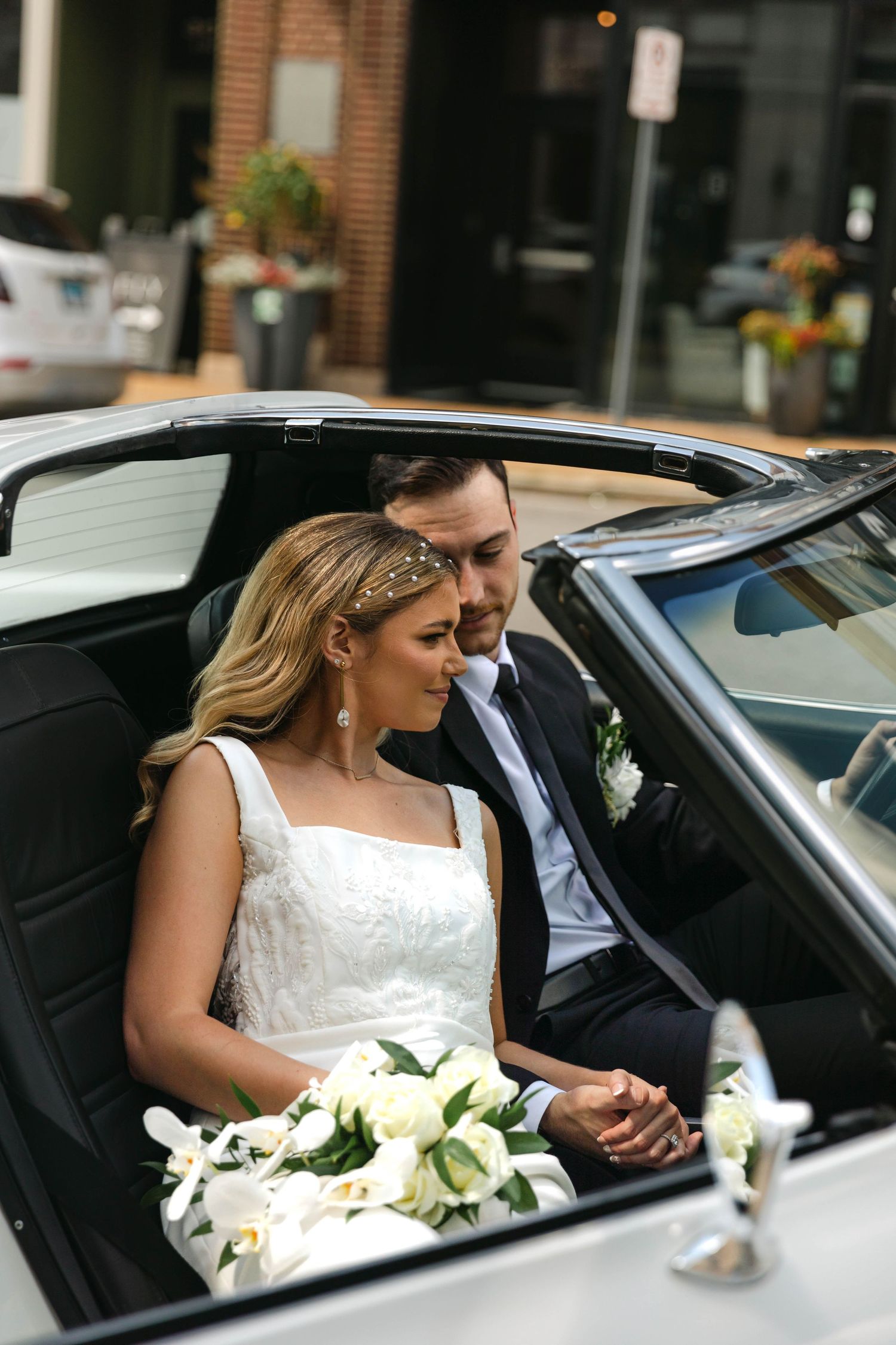 A couple shares a moment together in a black wedding car with white floral bouquet.