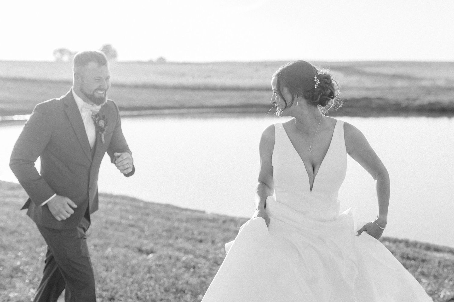 A couple in wedding attire runs playfully along a waterfront in a black and white photograph.