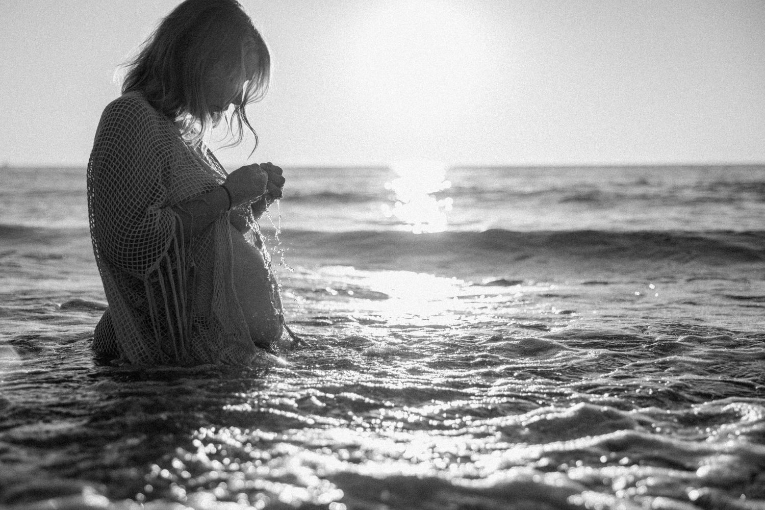 A maternity silhouette sits in shallow ocean waves at sunrise creating a moody black and white artistic scene during a maternity photoshoot