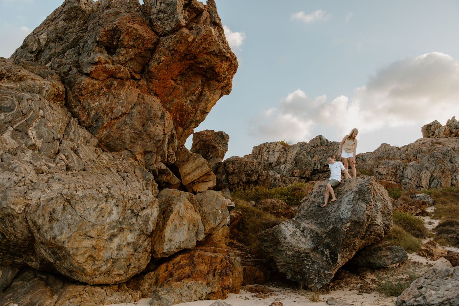 Two teens clamber over boulders at the beach during a sunset family photography session, In Point Samson, WA