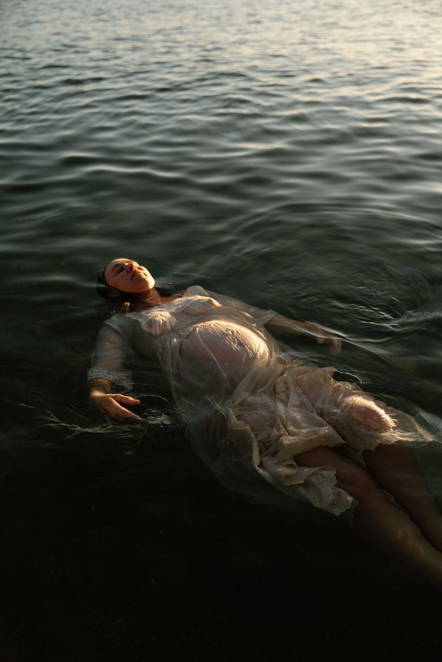 A figure in a white dress floats peacefully in dark rippling water during a sunrise maternity session in Wickham, WA