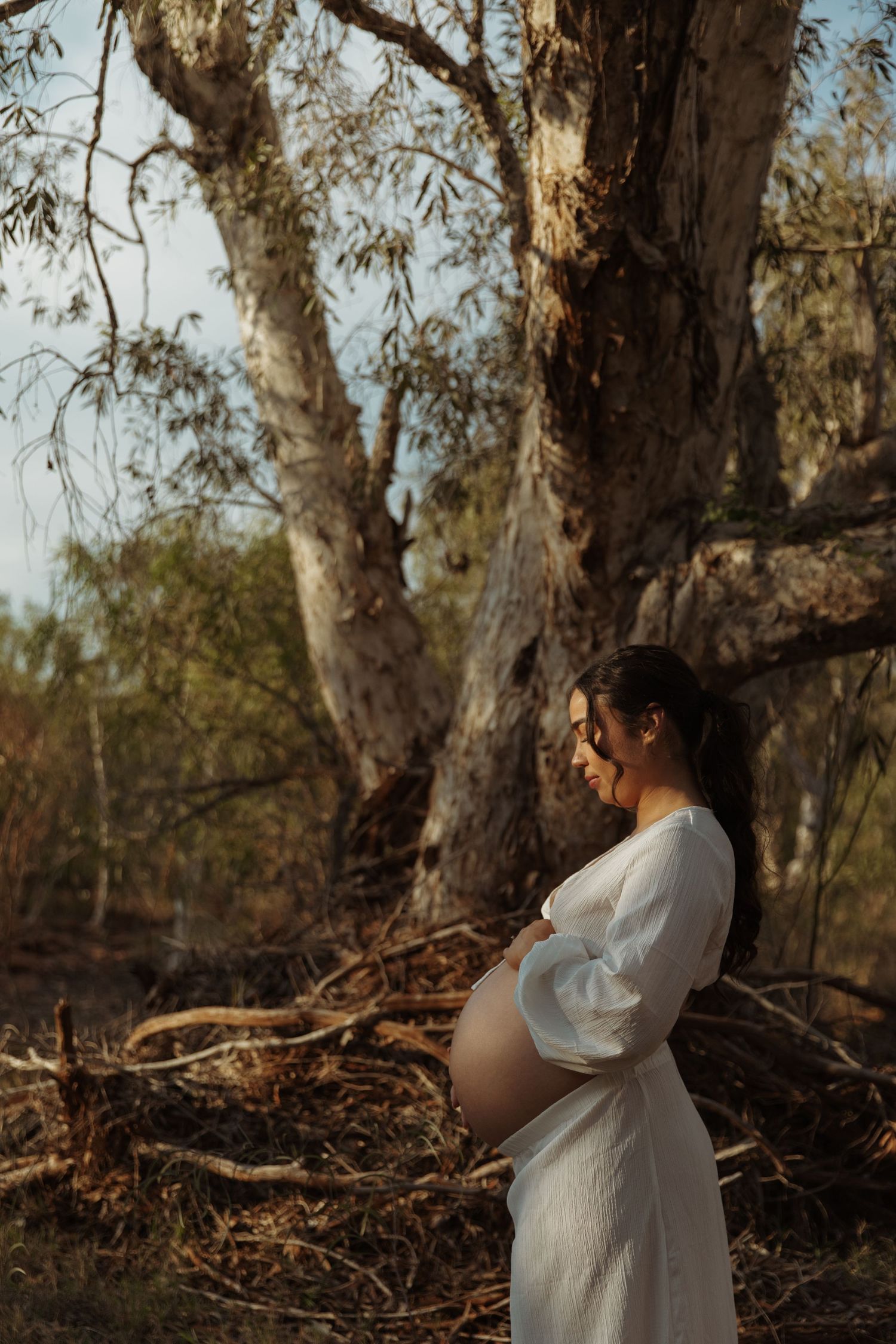 A pregnant woman in a white dress stands in a natural woodland setting during golden hour.