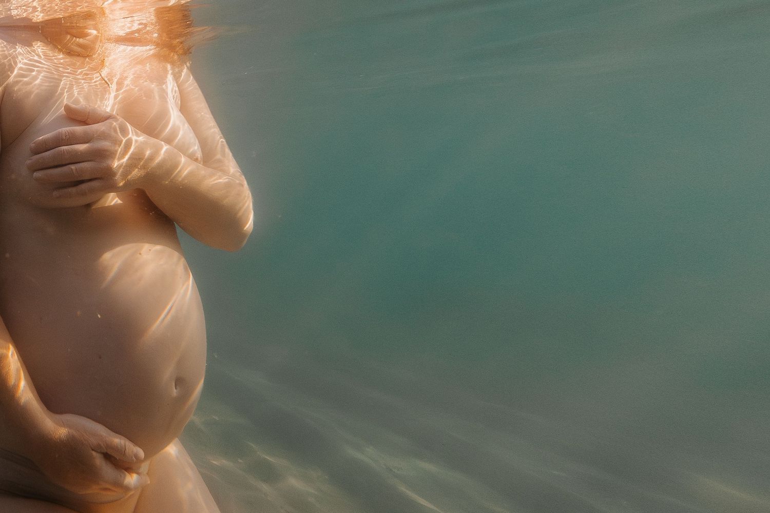 Underwater photo of pregnant belly in white fabric with sunlight filtering through turquoise water in Point Samson, WA