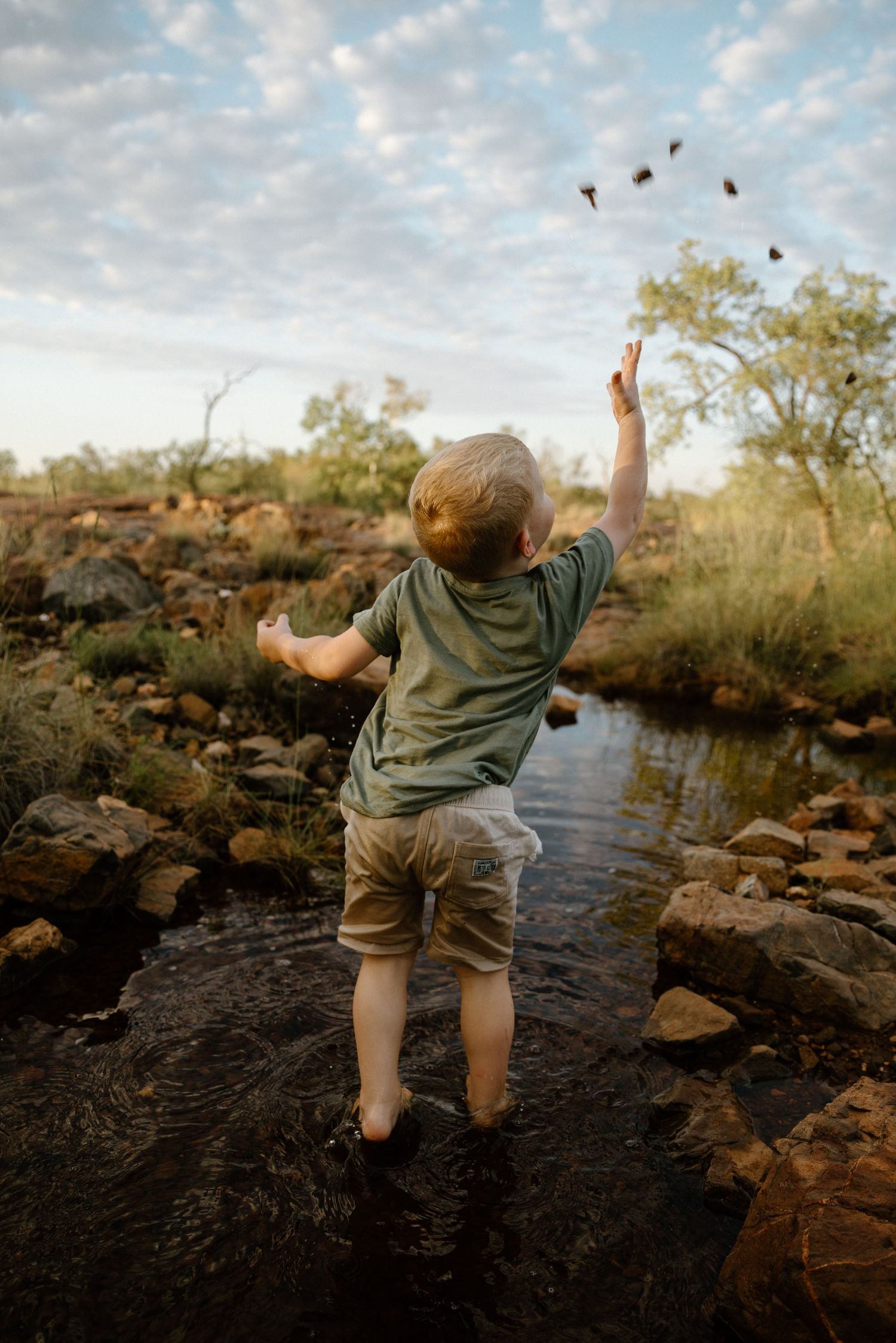 A young boy throws a handful of pebbles up in the air to land in the water during a motherhood photography session in Wickham, WA